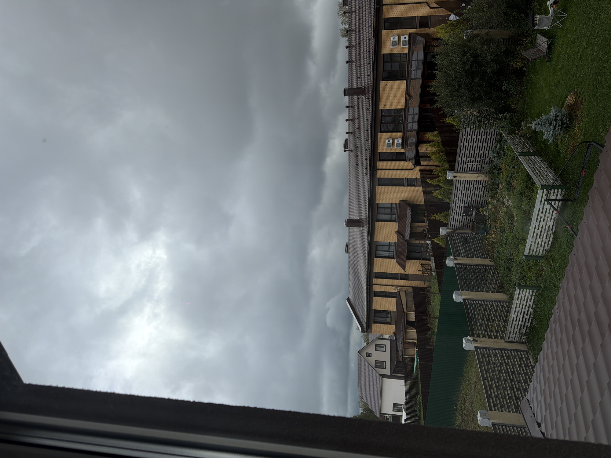 The image shows a view of overcast skies above a residential area. In the foreground, a brown tiled roof is visible. Beyond that, a backyard area is fenced with a dark green corrugated metal fence and a decorative wooden fence with concrete posts. The backyard contains overgrown vegetation, a small evergreen tree, two chairs, and a small table. In the middle ground, two townhouses are visible. The townhouse on the left is white with a dark roof and several windows. The townhouse on the right is made of yellow brick with a dark roof and multiple windows and air conditioning units. The sky is filled with dark, heavy clouds, suggesting impending rain or stormy weather. The scene appears to be a suburban or semi-rural setting.