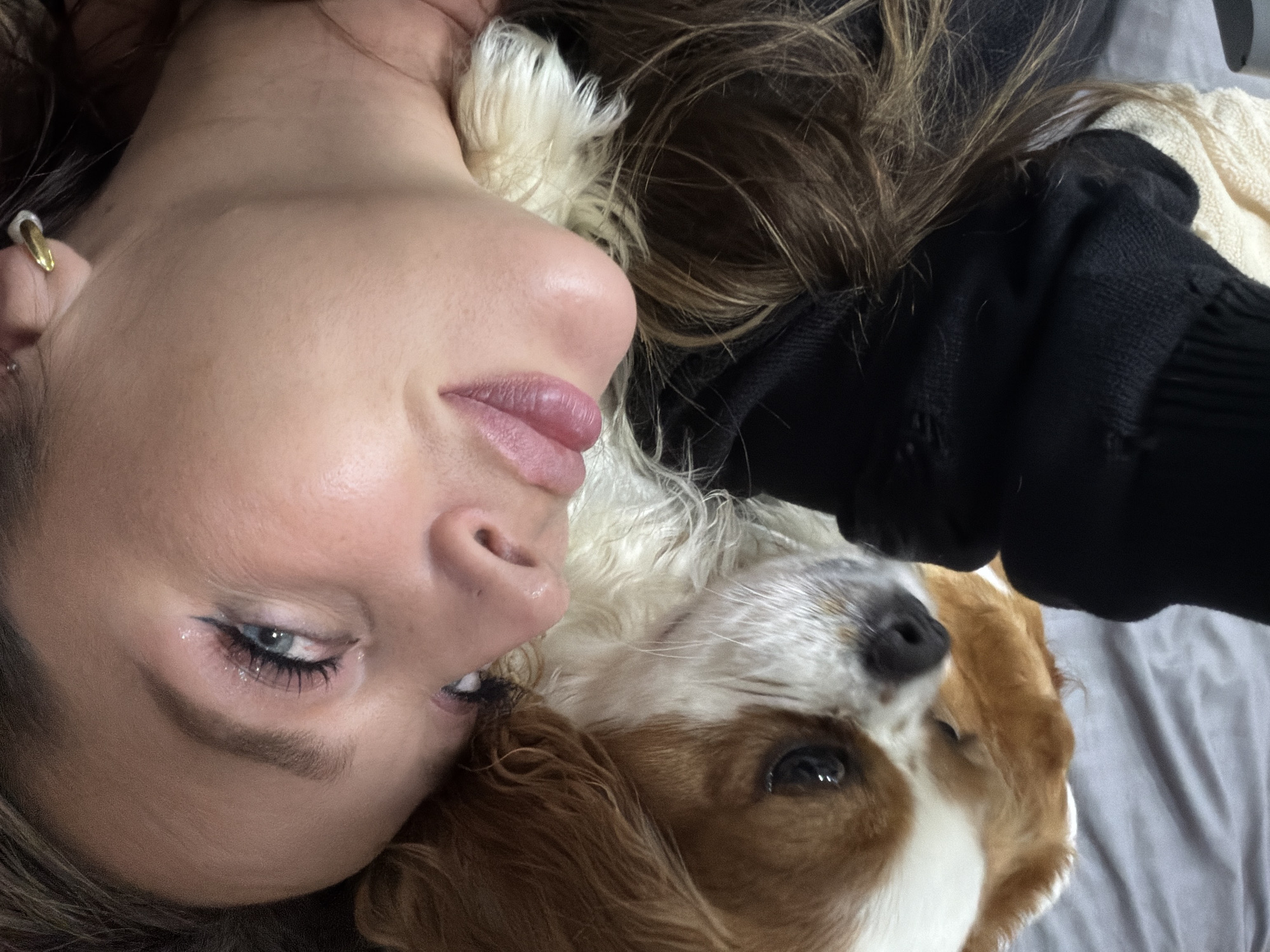A person and a dog are intimately resting together, seen from a tilted, slightly overhead perspective. The scene is set indoors, likely on a bed or sofa with grey bedding and a cream-colored blanket, in Chaikovsky, Russia.
The person, a young female, has fair skin, blue-grey eyes, and long, dark brown hair. She wears a golden hoop earring with a white element in her visible ear. Her makeup includes black eyeliner, mascara, and glossy, plump lips. She is wearing a dark, possibly black, long-sleeved top, part of which is visible on her shoulder and arm. Her expression is calm and relaxed.
Cuddled close to her head is a dog, appearing to be a Cavalier King Charles Spaniel or a similar breed, with soft white and light brown/tan fur. The dog's head is resting alongside the person's, and its dark eyes and nose are partially visible. Its fur appears soft and well-groomed.
The image captures a moment of quiet companionship and comfort between the two subjects. The lighting is soft and even, suggesting an indoor setting without harsh direct light. There is no visible text in the image.