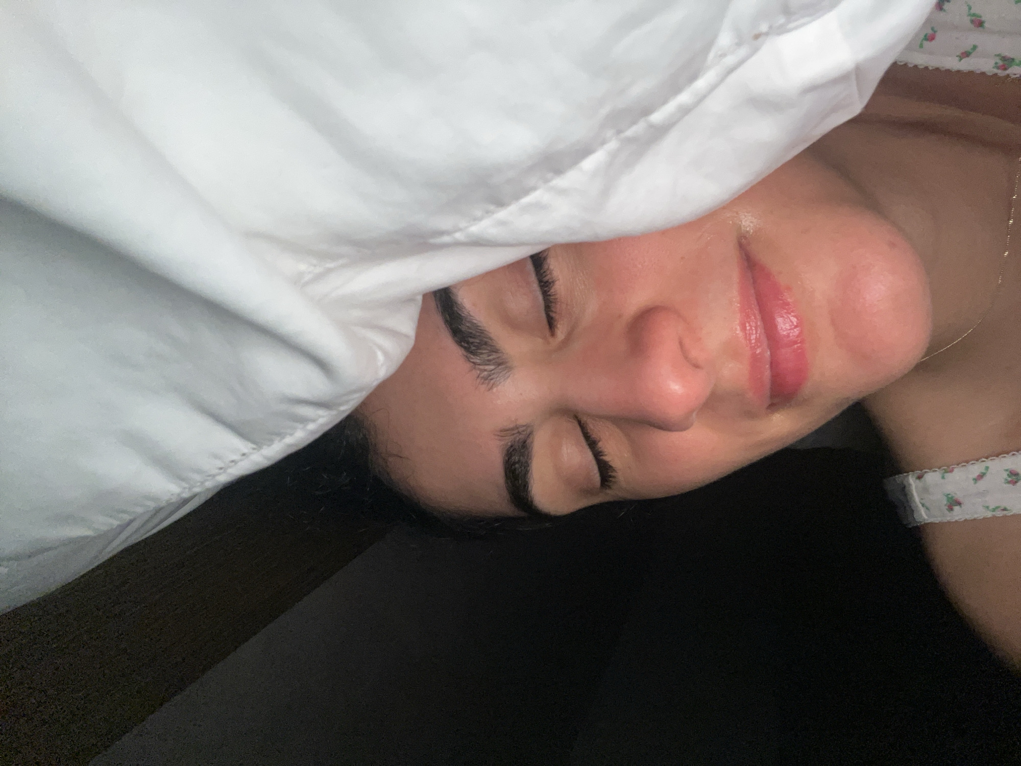 A person lies under a white pillow, face visible. The person's eyes are closed. A necklace is visible on their neck. The person wears a white floral-patterned top. The background is dark. It is not possible to ascertain the location of the image within Miami Beach, United States of America.