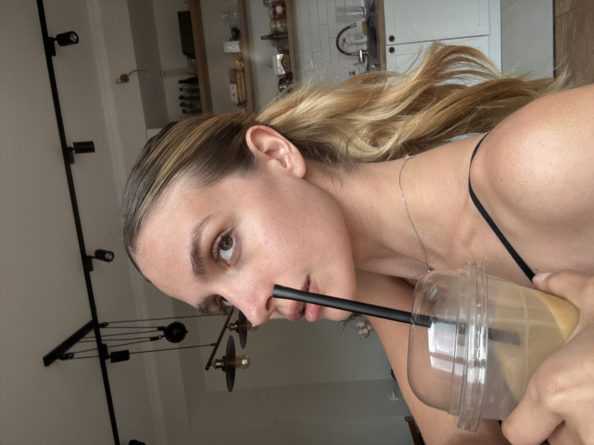A person with light brown hair pulled back into a ponytail is holding a clear plastic cup with a black straw, drinking from it. The person is wearing a thin black strap tank top and a delicate silver necklace. The background includes a kitchen with white cabinets and shelves stocked with various items. A black metal lighting fixture is also visible on the ceiling. The location appears to be indoors in an unknown city.