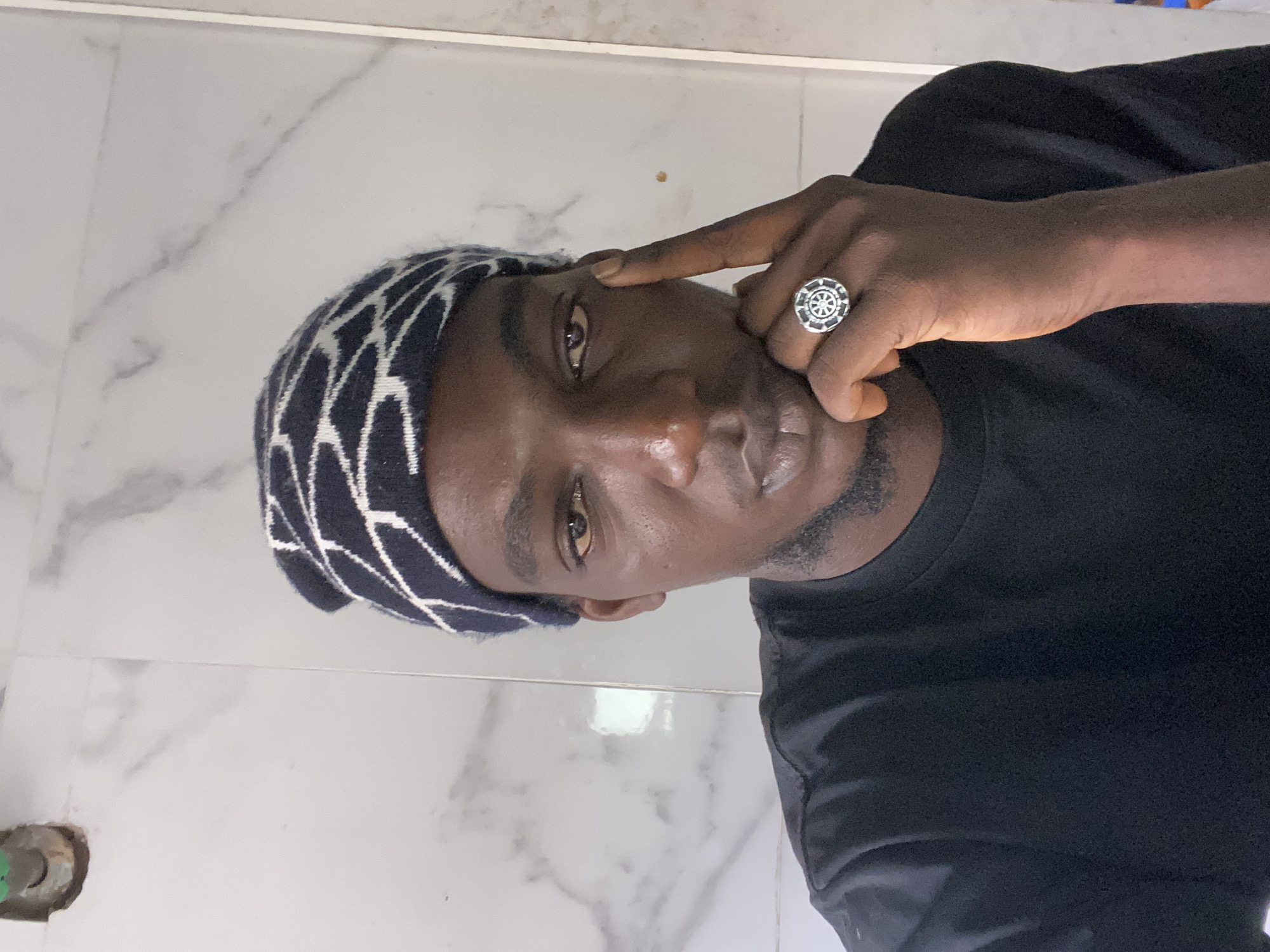 A dark-skinned male poses in a vertical selfie. He wears a navy blue and white patterned beanie and a black t-shirt. A silver ring adorns his right ring finger, which is near his right eyebrow. The background appears to be a marble-tiled surface. There are no definitive indicators within the image to confirm the location as Lagos, Nigeria.