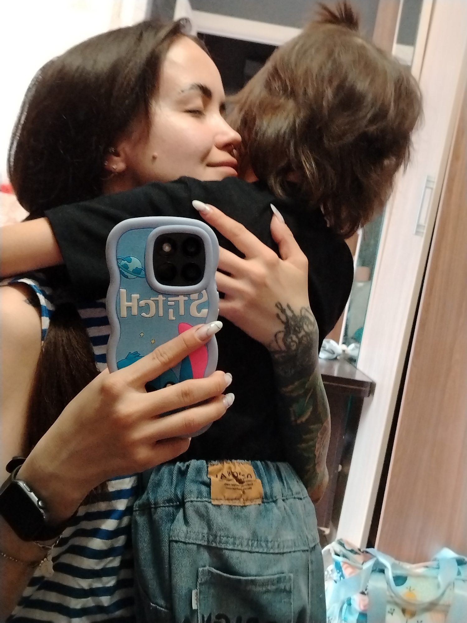A woman with long brown hair and a tattoo on her left arm embraces a small child with brown hair. She is holding a smartphone with a blue "Stitch" themed case. The woman is wearing a striped shirt and denim bottoms. They appear to be inside a home, possibly in Noviy, Russia, based on the image's metadata.