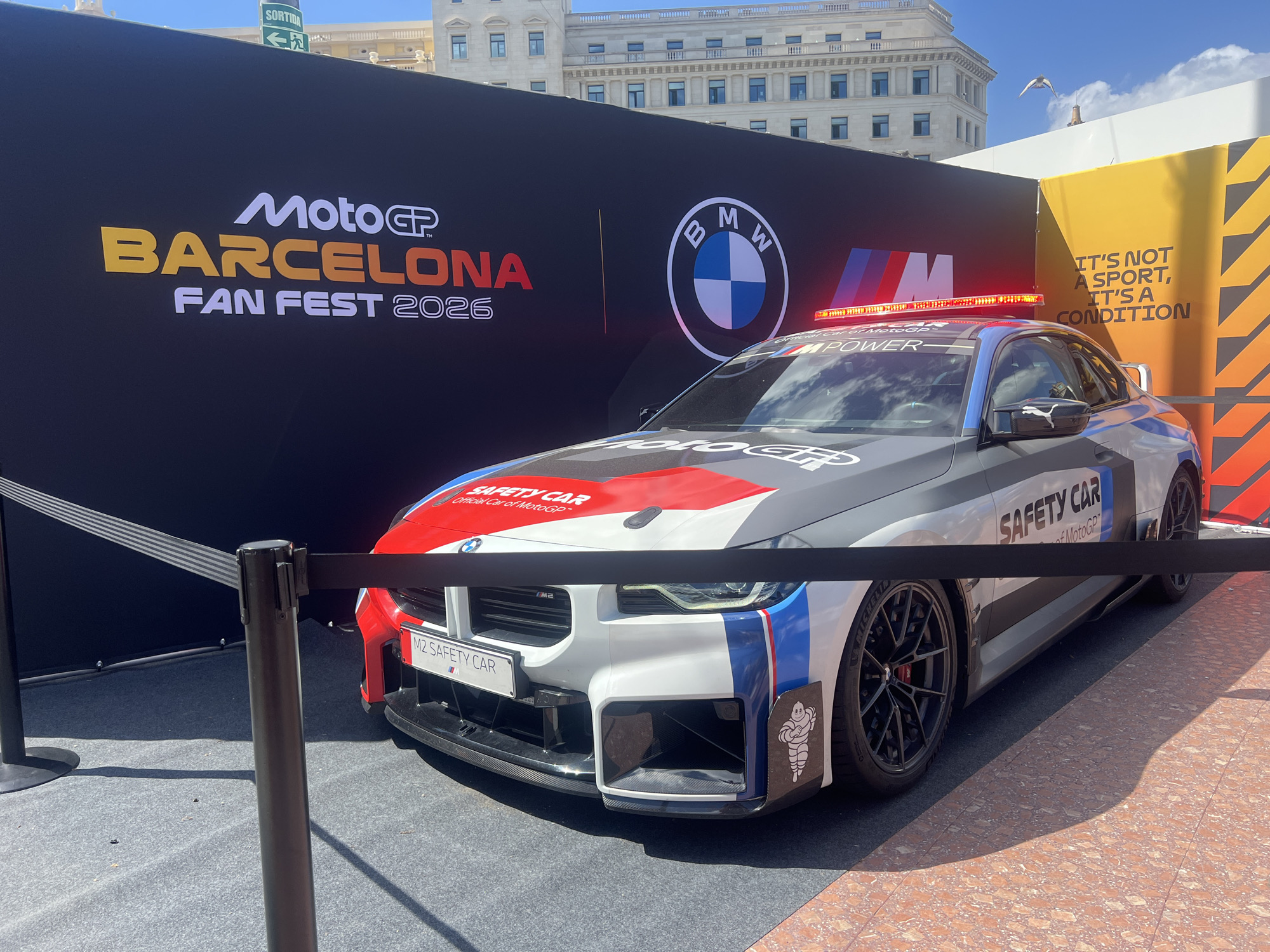 The image shows a BMW M2 Safety Car at the MotoGP Barcelona Fan Fest 2026 in Barcelona, Spain. The car has a grey, white, and red livery with "SAFETY CAR" written on the hood and "M POWER" on the windshield. It has a light bar on the roof. The car is cordoned off by black stanchions with retractable belts.
Behind the car is a black banner with "MotoGP BARCELONA FAN FEST 2026" and the BMW logo. To the right of the banner is a yellow sign with the text "IT'S NOT A SPORT, IT'S A CONDITION". In the background is a light-colored building with multiple windows. The sky is blue with a few white clouds. A bird is visible in the sky above the building.
