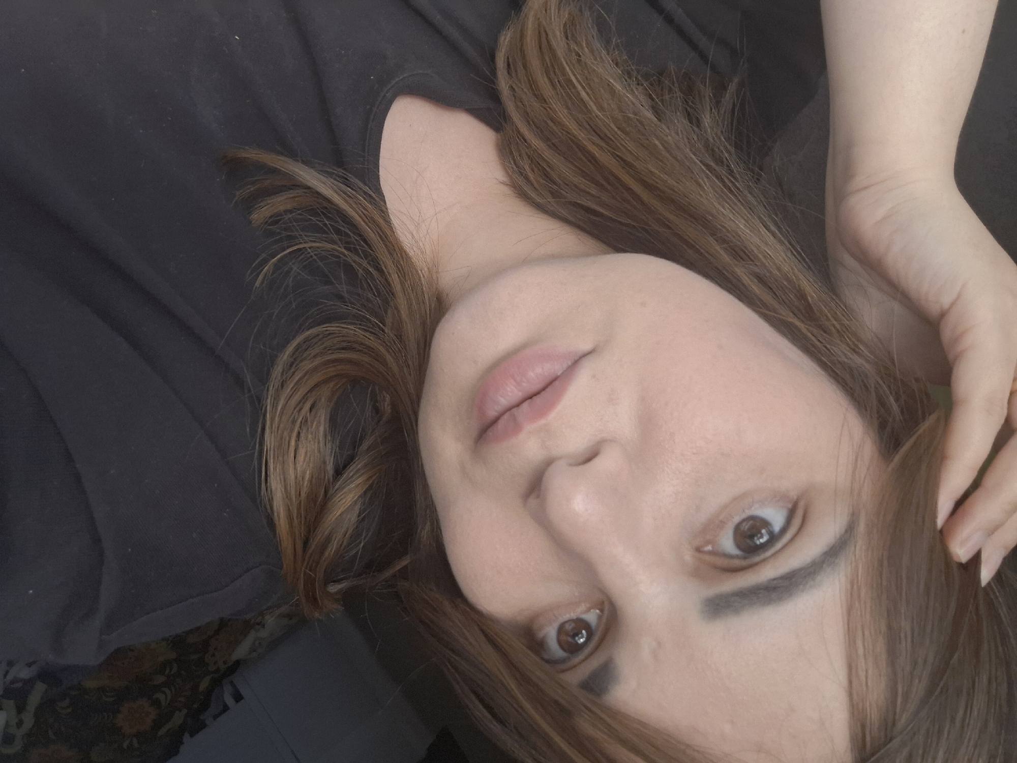 A medium close-up shot of a young woman is captured from a slightly elevated angle, her head tilted to her right. She has fair skin, brown eyes, and medium-length brown hair that is brushed to the side, with a few strands framing her face. Her left hand is raised, resting on her head, and she is wearing a black t-shirt. The background is dark and out of focus, with some patterned fabric visible on the right side. The image is taken indoors, likely in a residential setting, given the background details and the casual pose. There is no clear indication of a specific location within Chaikovsky, Russia.
