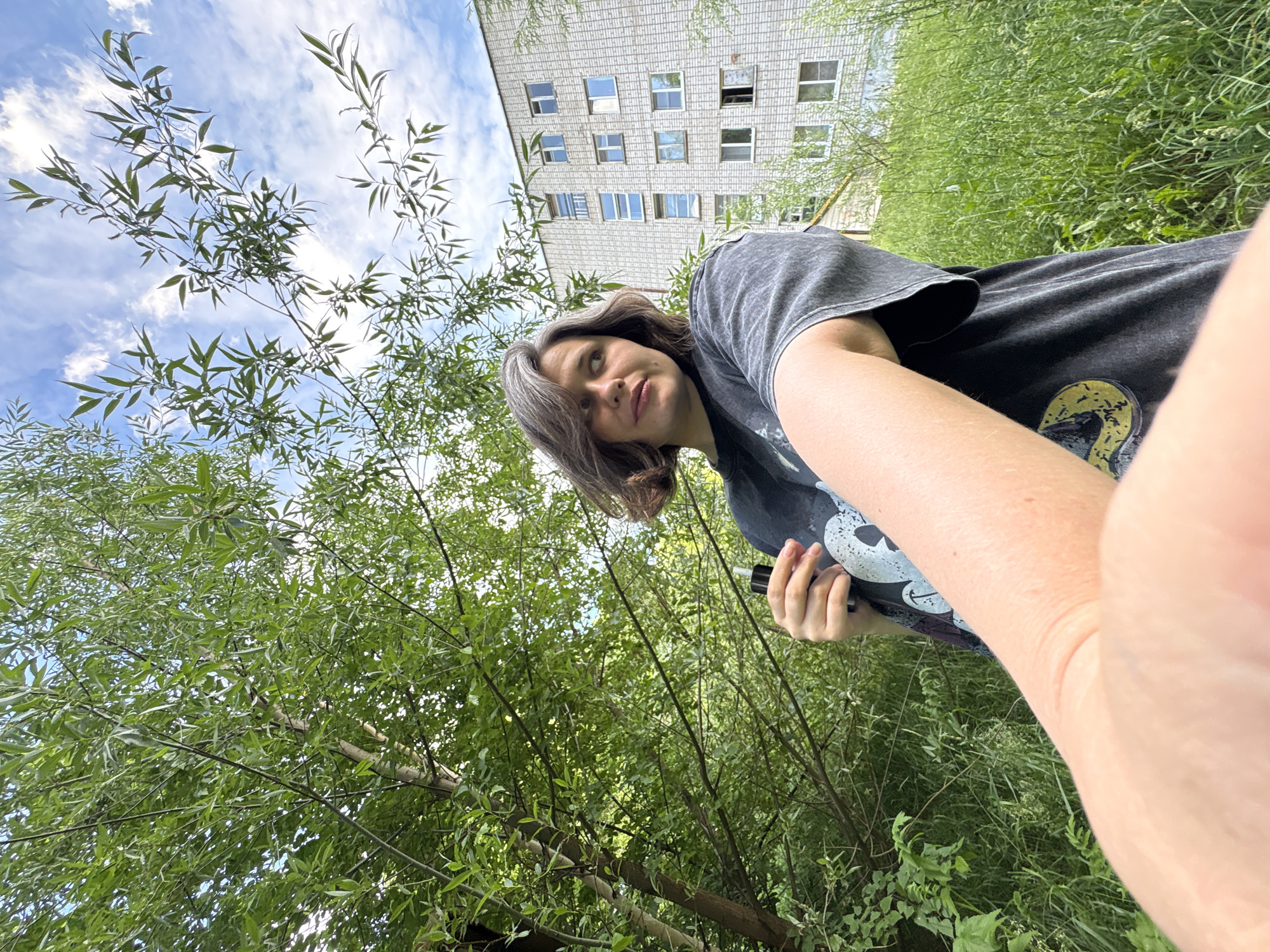A young woman with short, graying hair is taking a selfie outdoors. She is wearing a dark, oversized t-shirt with a graphic design. She holds a black electronic device in her right hand. The woman is looking away from the camera to her right. Behind her, a large tree with green leaves dominates the left side of the frame. In the background, a multi-story apartment building with a light-colored brick facade and numerous windows is visible. Tall green grass fills the ground area. The sky is blue with scattered white clouds. The scene appears to be located in a residential area of Chaikovsky, Russia, possibly a courtyard or a park adjacent to an apartment complex.