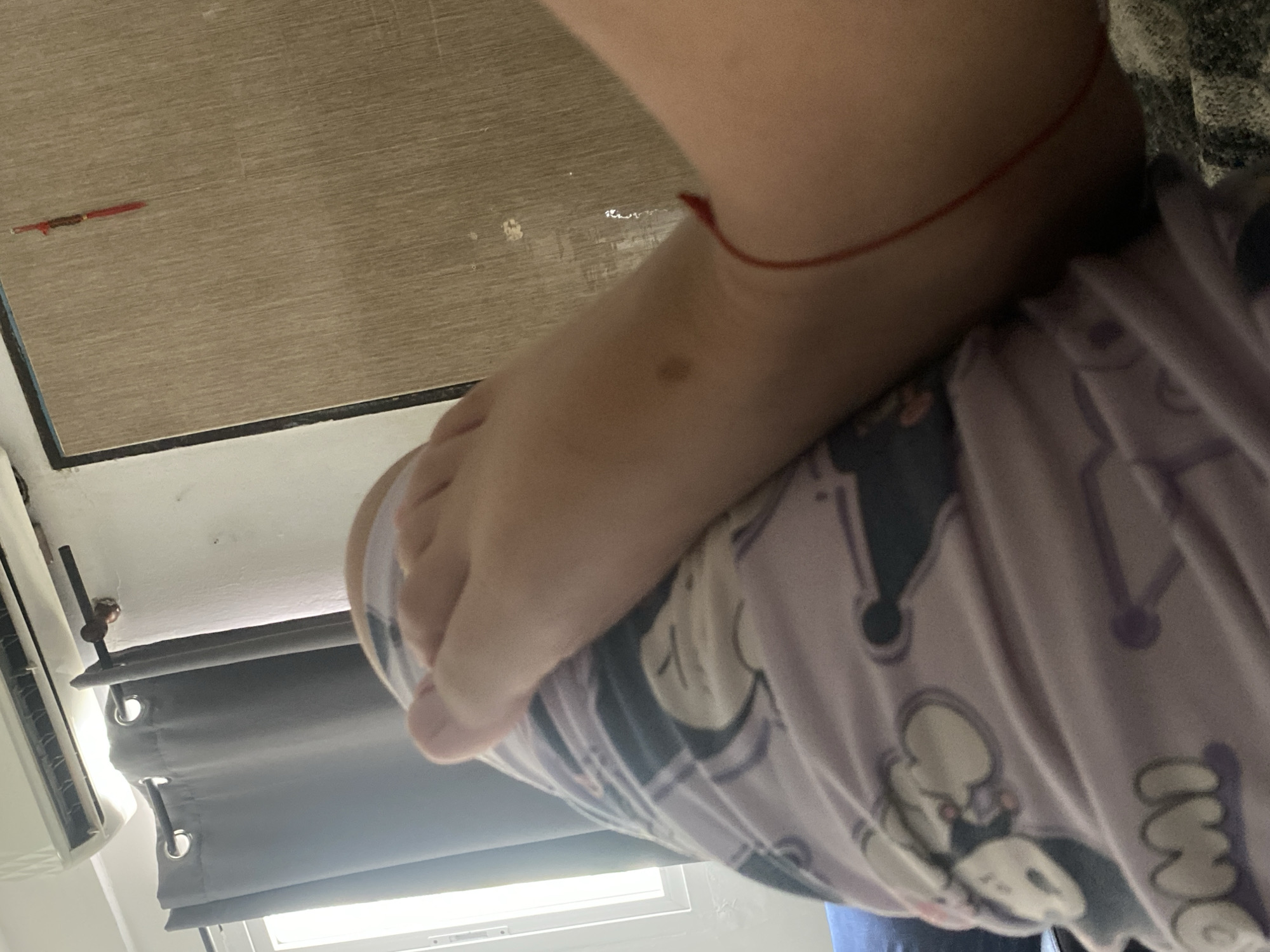 A close-up shot shows a foot resting on top of pajama pants with a cartoon print. The person is wearing a red string anklet. The background includes a wooden surface, a gray curtain, and what appears to be an air conditioning unit near a window. The location in Cotoperíz, Venezuela, is not discernable from the image.