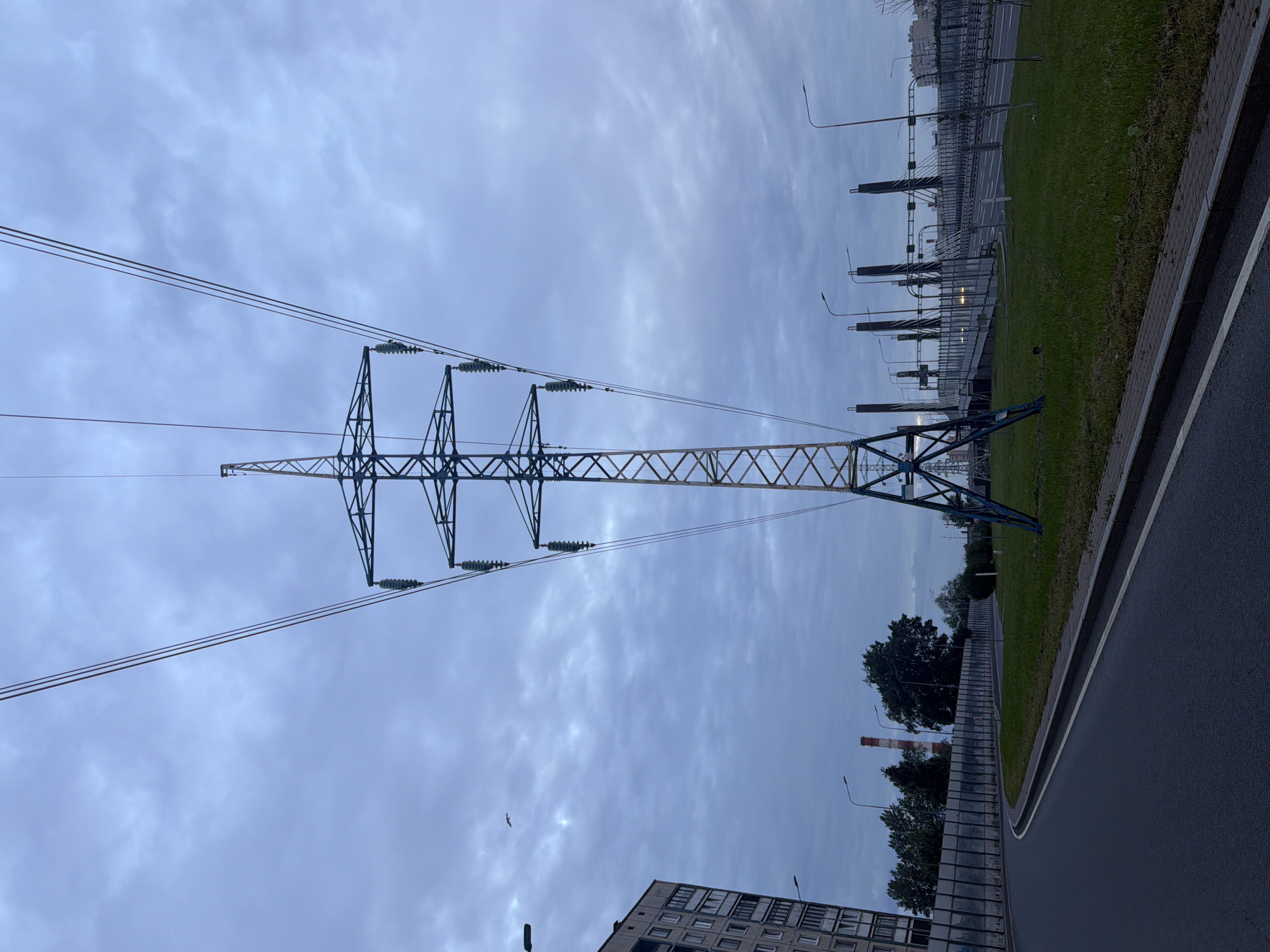An outdoor, eye-level, full shot captures a large electric transmission tower with a dark blue and yellow lattice structure in a city. The tower is positioned centrally and dominates the frame, with thick power lines extending outwards from its apex into the cloudy, overcast sky. To the left, a multi-story building with visible windows and balconies is partially visible. A paved road curves in the foreground, bordered by a grassy median and a noise barrier fence. Beyond the fence, there are trees and industrial structures, including a red and white smokestack. In the distance, a bridge with distinctive pylons and overhead lights can be seen. The lighting suggests it is either early morning or late evening.