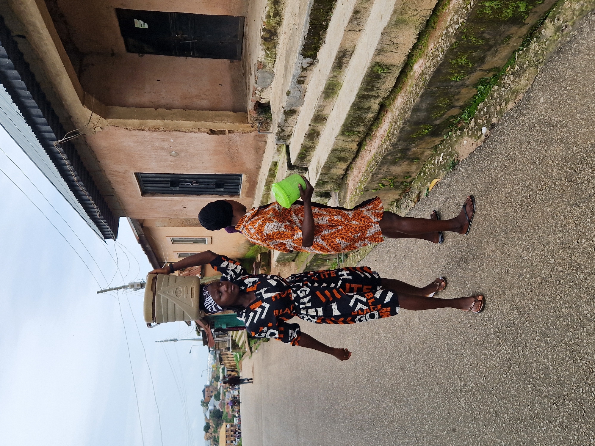 In Kabong, Nigeria, two young women are depicted walking on an asphalt road. The woman on the left carries a large, light-colored plastic container on her head, balanced with a patterned cloth. She is wearing a short, black dress with a white and orange geometric pattern. The woman on the right walks beside her, holding a green plastic container. She is wearing a short, orange and white patterned dress and a dark head covering. They are walking away from the camera, on the right side of the road. In the background, there are several buildings with tan walls and corrugated metal roofs. The architecture suggests a residential or commercial area. The sky is overcast. The environment appears to be a street scene in a developing urban area, with visible overhead electrical wires and utility poles. The ground in the foreground and on the side of the road is rough and textured, with some vegetation growing along the concrete drainage channels.