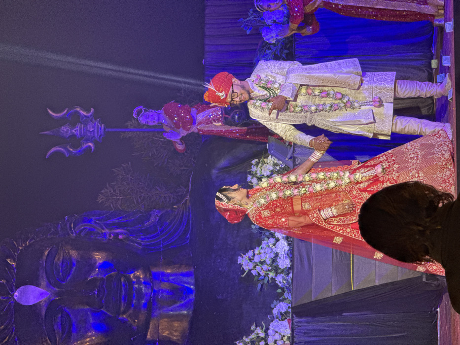 A wedding scene shows a bride and groom in traditional Indian attire. The bride wears a red lehenga with gold embroidery and a garland of roses. The groom is in a cream-colored sherwani with a matching garland. They hold hands and appear to be dancing. In the background, a large, dark sculpture of Shiva's face is visible, along with a trident and a statue of a goddess. The scene is illuminated with blue and white lights.
