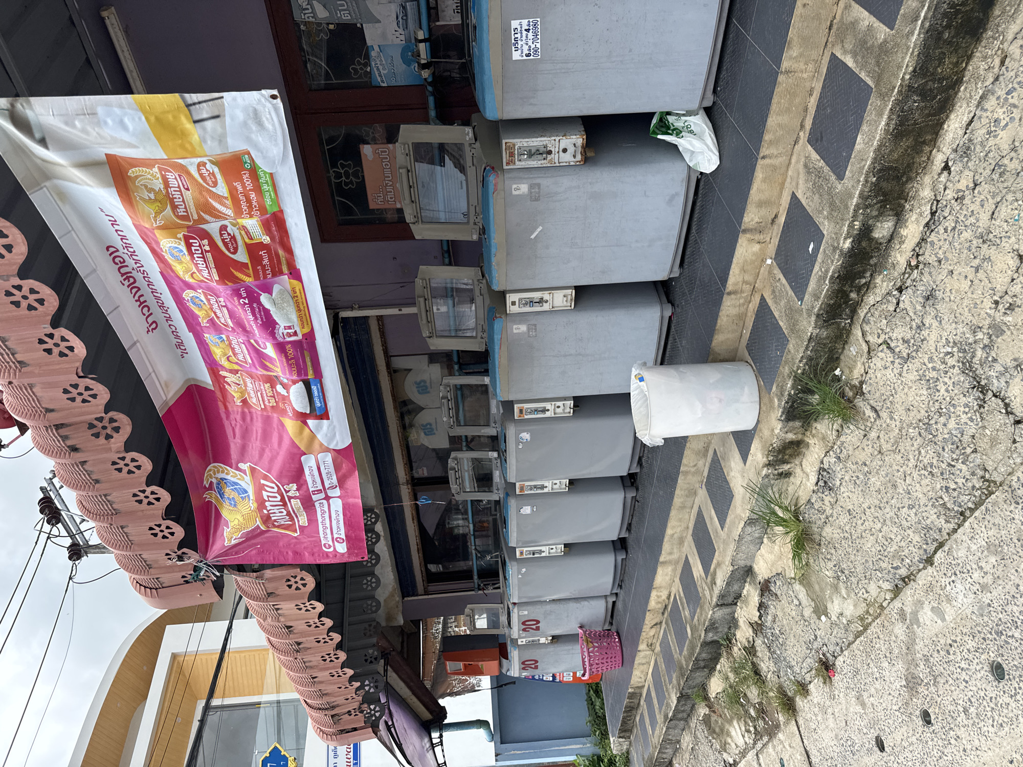 A row of commercial washing machines is parked outside a building on a street in Rawai, Thailand. The washing machines are grey with blue accents and are arranged in a line on a raised platform in front of the building. Some of the machines have labels with Thai writing and numbers. A pink laundry basket is placed on the ground in front of the first washing machine, and a white trash can is next to it. A banner with Thai writing and images of rice packages is hanging above the washing machines. The building behind the machines appears to be a shop or commercial establishment with windows and a dark-colored awning. The street in front of the building is paved, and there is some greenery growing in cracks in the pavement. The sky is overcast, suggesting cloudy weather.