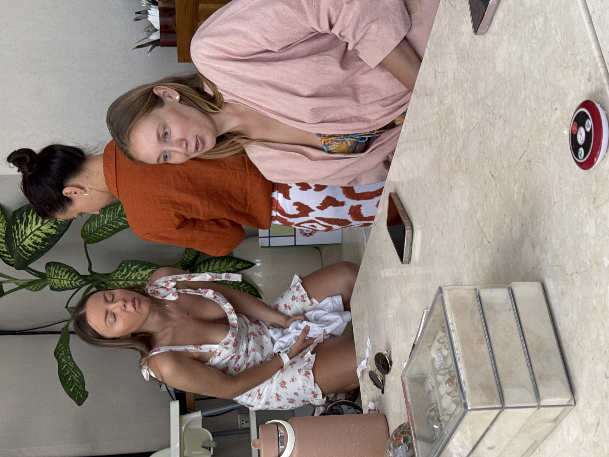 Three women are sitting at a marble table in an outdoor setting, presumably in a cafe. The woman closest to the camera is wearing a white dress with a floral pattern. She is sitting down, looking down, and holding something white in her lap. The second woman wears an orange blouse and is leaning in, looking at the woman in the floral dress. The third woman is to the right of the second woman; she wears a pink shirt and is also looking towards the woman in the floral dress. The table has two mobile phones and a restaurant paging device on it. There's a jewelry display box on the table, as well as some sunglasses. There are green plants nearby. The image suggests a candid moment between the women during a casual outing. The location is Ungasan, Indonesia.