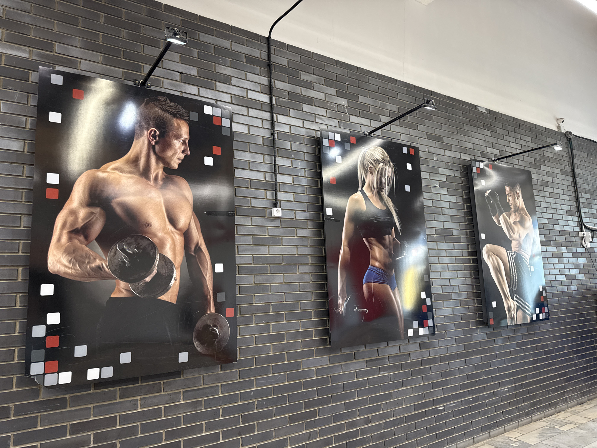 The image displays a dark brick wall adorned with three large, vertically oriented posters featuring muscular individuals engaged in fitness activities. The posters are illuminated by adjustable track lighting. The first poster on the left shows a shirtless man lifting a dumbbell, his physique highlighted by the lighting. The middle poster features a woman in athletic wear, also holding a dumbbell. The third poster, on the right, depicts a man in boxing shorts, with his body tensed as if in mid-fight. The posters have a decorative border of small squares in red, white, and black. Electrical conduits and a power outlet are visible on the wall, along with a security camera in the upper right corner. The overall scene suggests a gym or a fitness-related establishment. The location is identified as Chaikovsky, Russia.