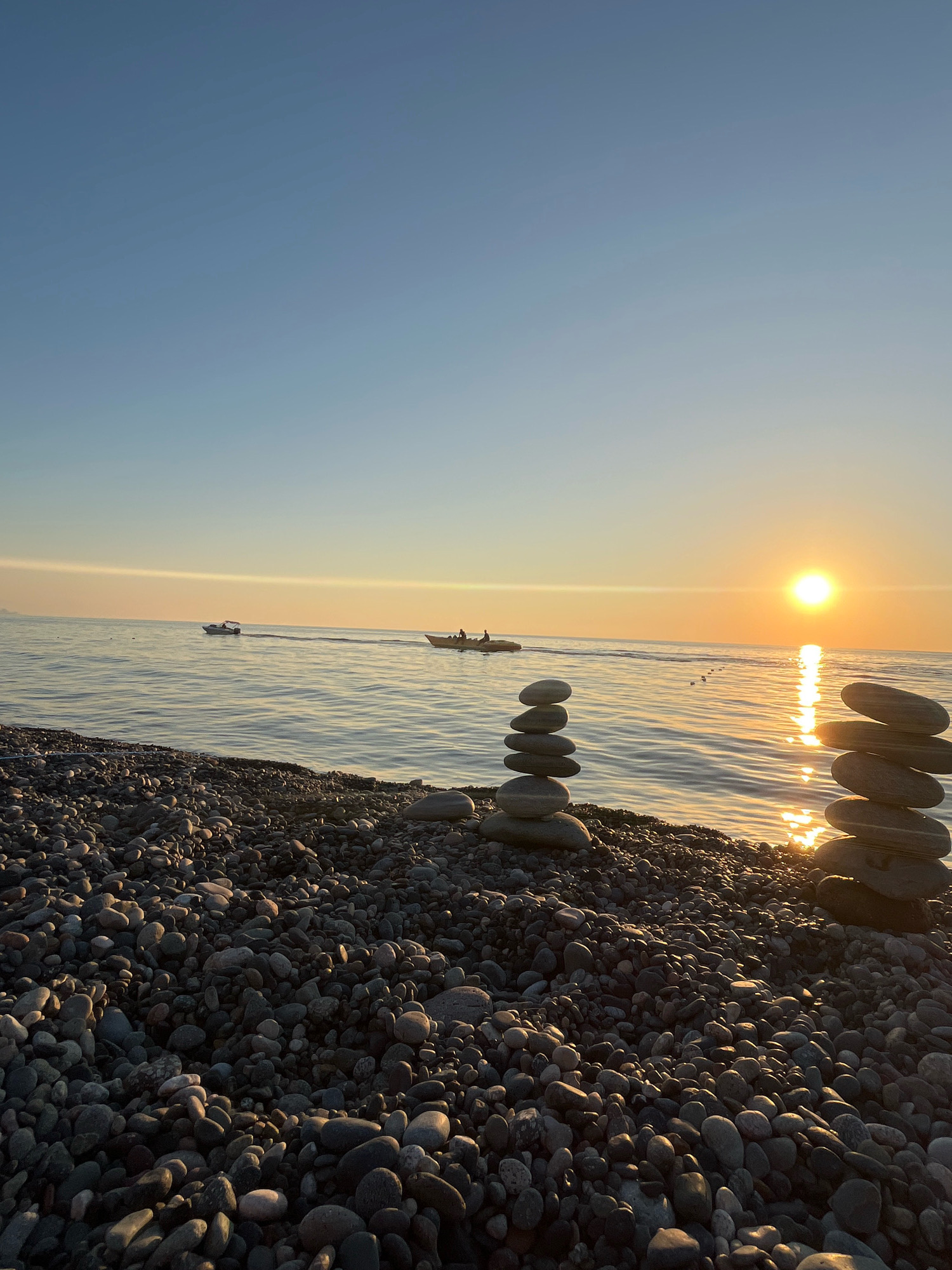 The image depicts a serene sunset scene on a pebble beach in Kobuleti, Georgia. In the foreground, two stacks of smooth, grey stones are artfully balanced on the shore. The sea stretches out to the horizon, reflecting the warm glow of the setting sun, which is positioned slightly off-center to the right. The sky transitions from a clear, soft blue at the top to a gradient of warm oranges and yellows near the horizon. Two small boats are visible on the water; one is further out, while another is closer to the shore, with figures visible onboard. The beach is covered in a dense layer of pebbles, with some larger stones scattered throughout. The overall atmosphere is peaceful and tranquil, with the setting sun casting a golden light over the water and the coastline.