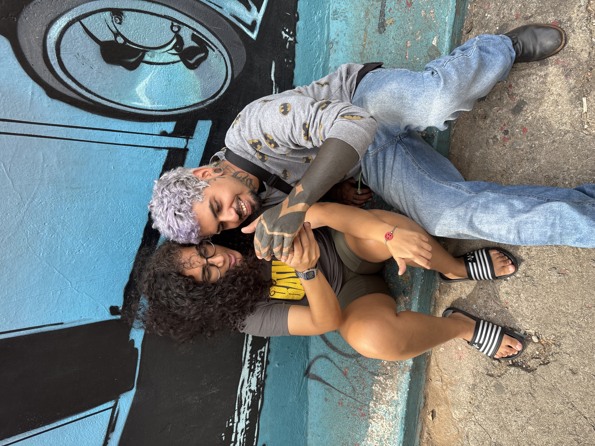 Two young adults, a man and a woman, are lying on a concrete surface next to a wall covered in graffiti. The man is positioned slightly above the woman, their bodies angled. He has light purple hair, facial piercings, and a tattoo that covers his forearm and hand in solid black. He wears a grey sweatshirt patterned with black Batman symbols and light blue jeans, and a black boot is visible at the edge of the image. The woman has dark curly hair and is wearing glasses, a dark t-shirt, and olive green shorts. A watch is visible on her left wrist and a bracelet on her right. She wears Adidas slide sandals. Both individuals are smiling and holding hands. The graffiti on the wall includes a large, stylized drawing of a tire and other abstract shapes in black and shades of blue. The concrete surface is textured and has some discoloration. The image is taken in daylight with no evidence that it is located in the city of Sao Paulo, Brazil.