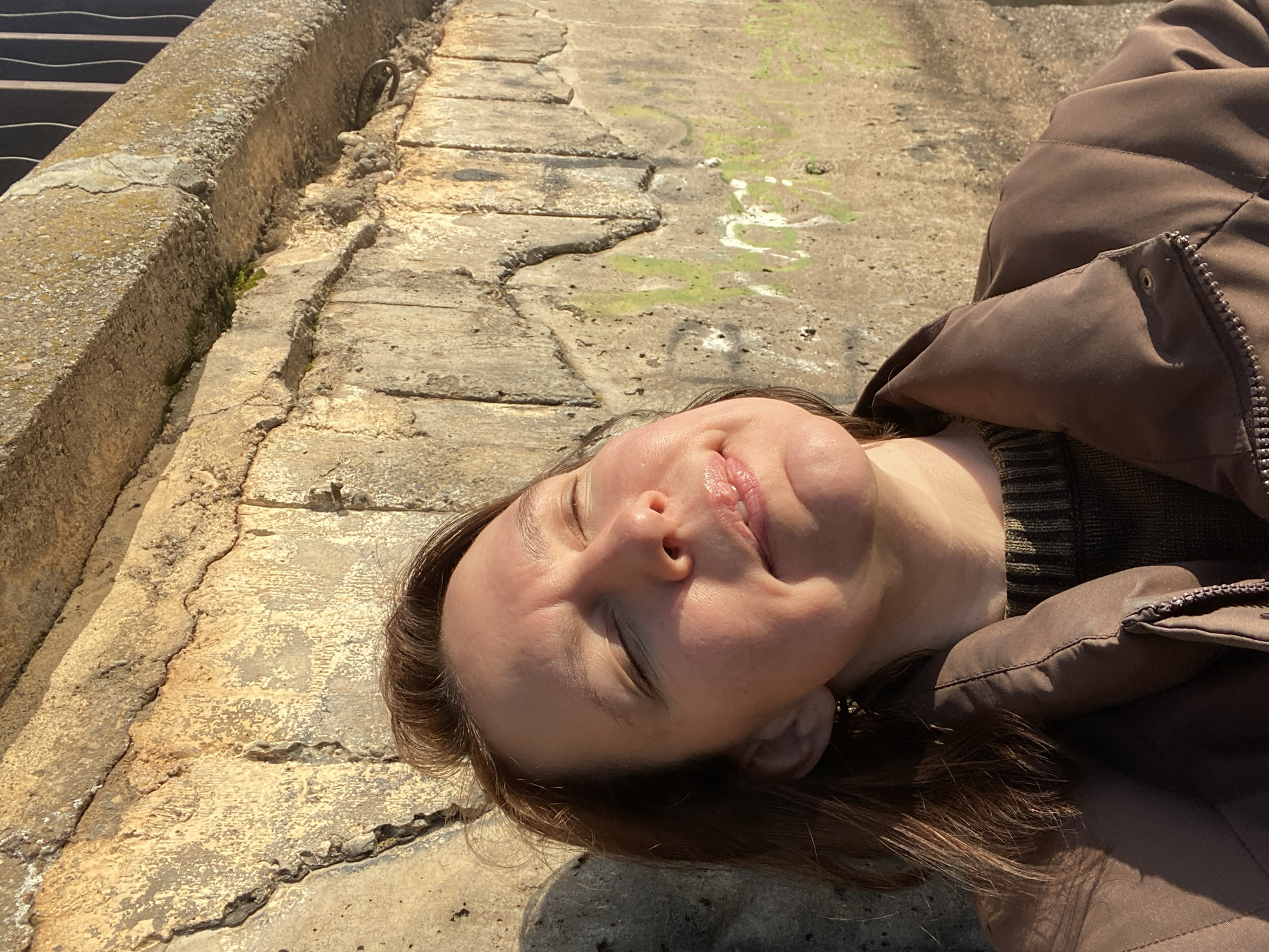 A woman is depicted lying on her back on a rough, cracked concrete surface, looking upwards with her eyes gently closed and a serene expression. Her dark brown hair spreads around her head. She is wearing a brown puffer jacket over a dark, possibly striped, sweater. Bright sunlight casts strong shadows, indicating a clear, sunny day. To her left, a weathered concrete curb or wall runs along the edge of the scene, with a glimpse of a metal grate partially visible. The ground exhibits signs of age and neglect, with visible cracks, uneven textures, and patches of green moss or algae. The setting appears to be an outdoor, utilitarian area in Chaikovsky, Russia. No text is visible in the image.