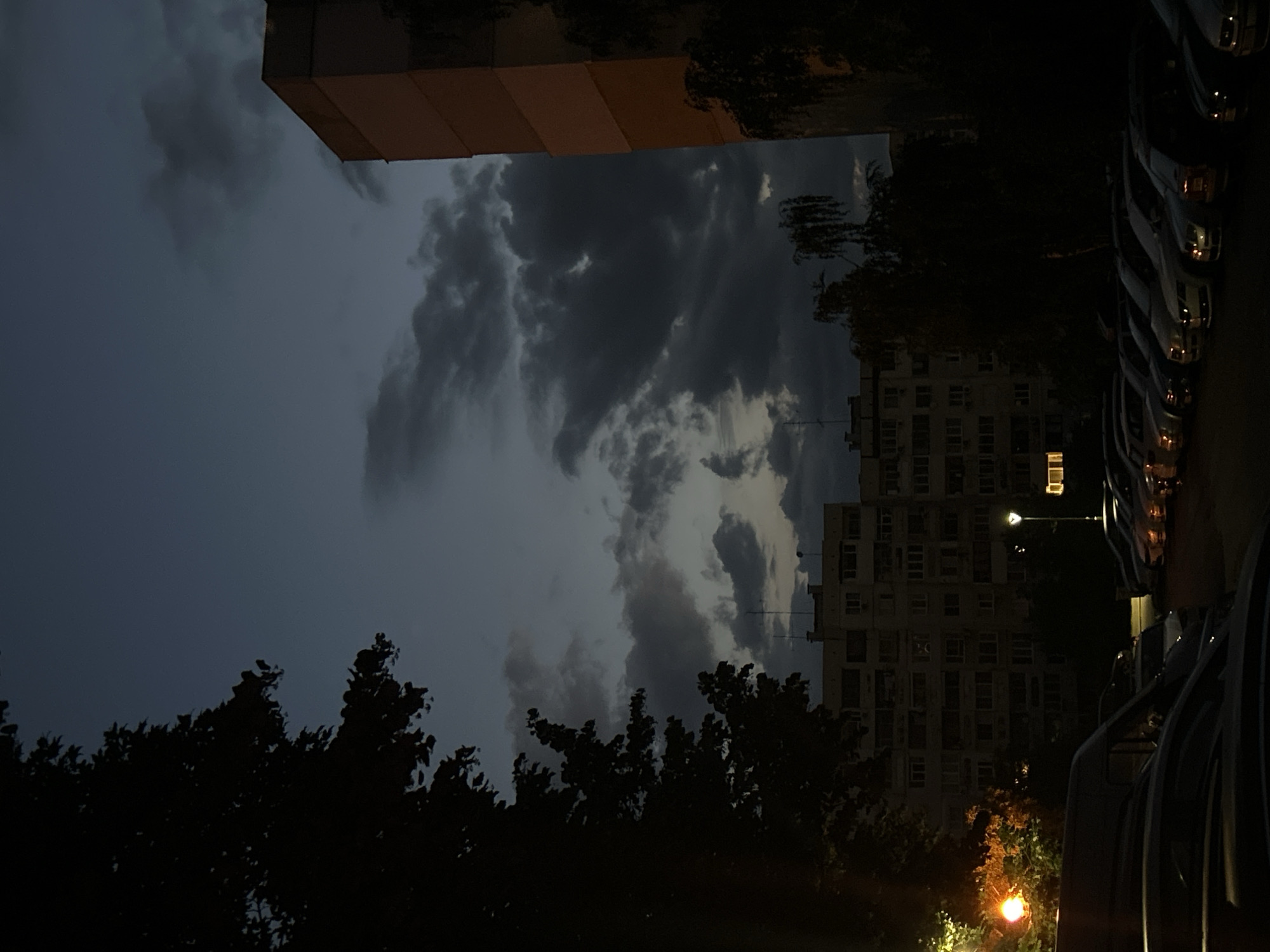 The image depicts a nighttime or dusk scene in a residential area, likely a city. The sky is a dark, moody blue with prominent, shadowy clouds, suggesting it is either late evening or overcast. Silhouetted trees frame the left side of the image, their foliage dense and dark.
In the mid-ground, several apartment buildings are visible. The central building is a multi-story structure with a beige facade and numerous windows, some of which are lit, indicating occupancy. To the right, a taller building with an orange-toned facade is partially visible, its upper sections obscured by the darkness and potentially more trees.
In the foreground, a parking lot is filled with numerous cars parked in rows. The vehicles are mostly dark-colored, and their outlines are softly illuminated by ambient light, possibly streetlights or light spilling from the buildings. A streetlight with a visible globe emits a warm orange glow on the left side of the frame, near a patch of illuminated foliage. Another streetlight with a white glow is visible further back in the parking lot. The overall impression is one of a quiet, urban residential scene as day transitions into night.