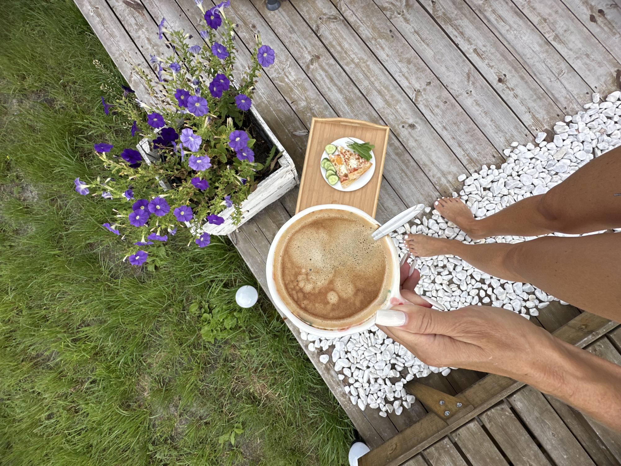 A first-person perspective shows a person's hand holding a white mug of coffee with a frothy top. The mug has a subtle floral pattern. The person's feet, bare with white painted toenails, are visible, standing on a wooden deck. The deck is partially covered with small white pebbles. Adjacent to the coffee mug is a small wooden tray holding a slice of pizza, cucumber slices, and a green garnish. In the background, a white planter is filled with vibrant purple petunias. To the left of the deck, a patch of green grass is visible, with two white spheres resembling golf balls on the ground. The scene appears to be outdoors on a warm day. The location is not specified as Tyumen, Russia.