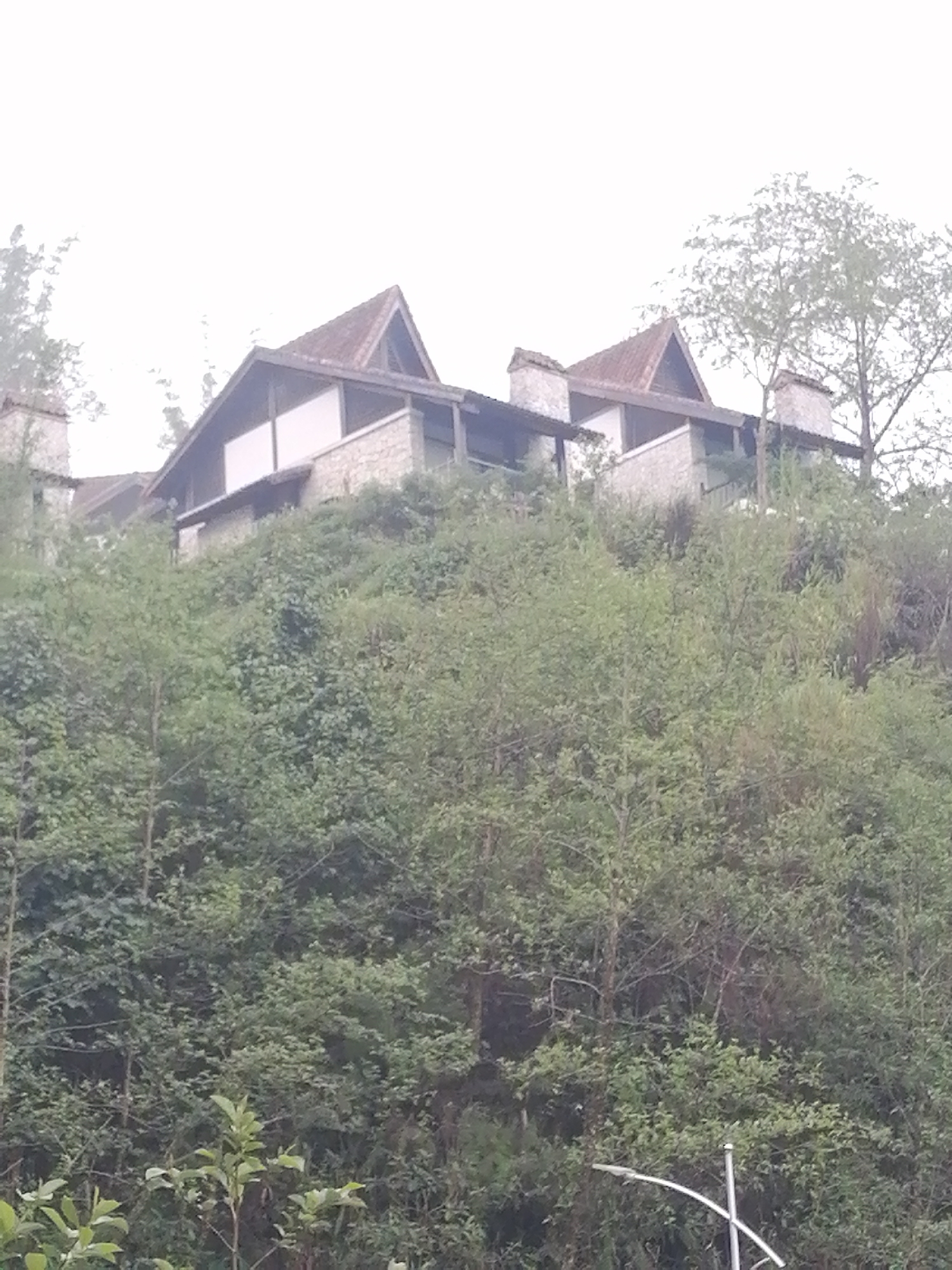 The image depicts a hillside in Sa Pa Ward, Vietnam, densely covered with trees and foliage. Atop the hill, several buildings with distinct architectural features are visible. The buildings feature light-colored walls, dark roofing, and chimney-like structures. The sky is overcast, suggesting a cloudy day. The scene is shot from a low angle, emphasizing the elevated position of the buildings relative to the foreground vegetation. A partial view of a lamppost appears in the bottom right corner of the image.
