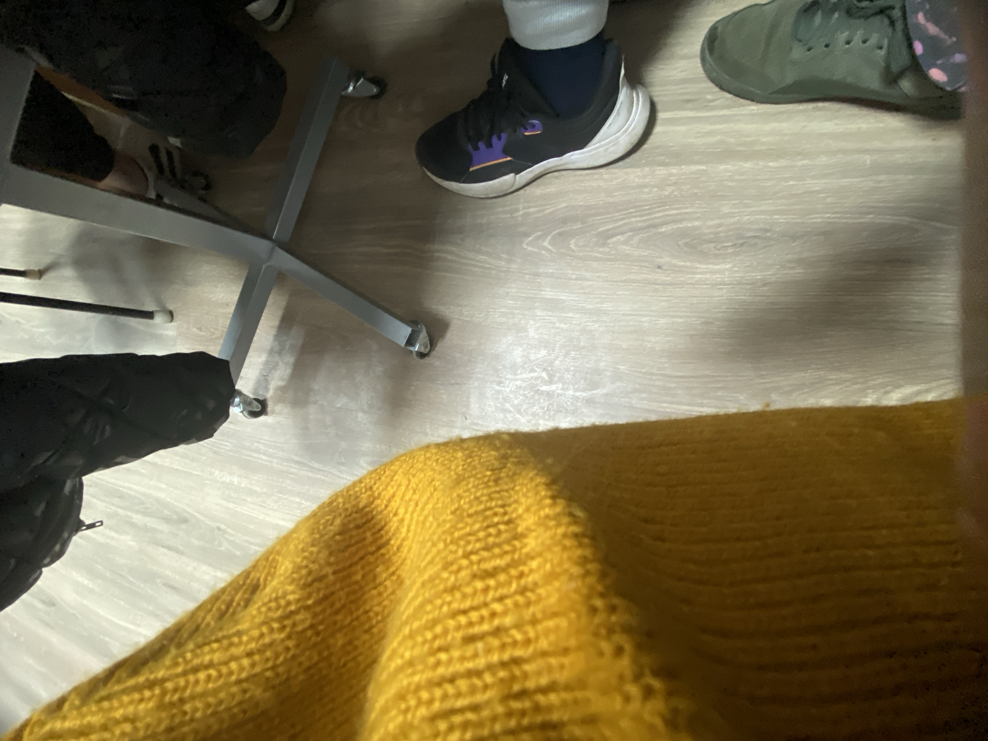 The image depicts a section of flooring with three pairs of shoes visible: a pair of dark sneakers with white soles and purple accents, a pair of green sneakers, and a pair of black shoes. A portion of a yellow knitted garment or blanket is in the foreground, partially obscuring the scene. The legs of a desk chair are also visible. There is no information in the image to identify the location as Troyan, Bulgaria.