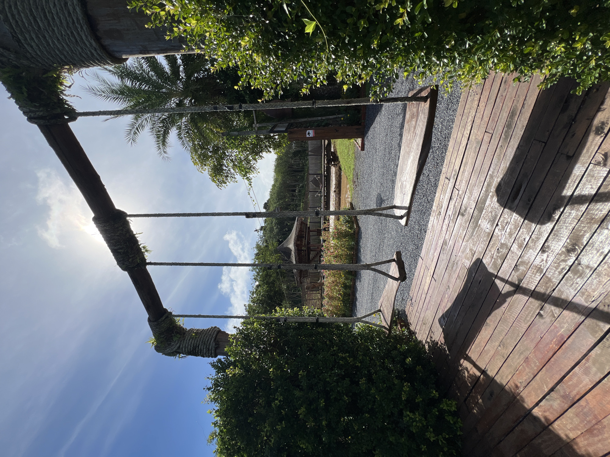 This image shows a swing set made of natural materials, including thick ropes and wooden beams. The swing set is set against a backdrop of lush green foliage and a bright blue sky with scattered clouds. In the distance, there are palm trees and a hint of a building with a thatched roof. The ground around the swing set is covered in dark gravel, and there's a wooden deck to the right of the frame. The sun is shining brightly, casting long shadows on the wooden deck, suggesting it is daytime, possibly late morning or early afternoon. The scene appears to be set in a tropical or subtropical location. There is no visible activity or people in the image, giving a sense of tranquility and a quiet, natural setting. A small sign with an icon is visible on a wooden post in the background, but the text is not legible. The overall impression is one of a peaceful outdoor space, perhaps a park or a resort, in Thailand.