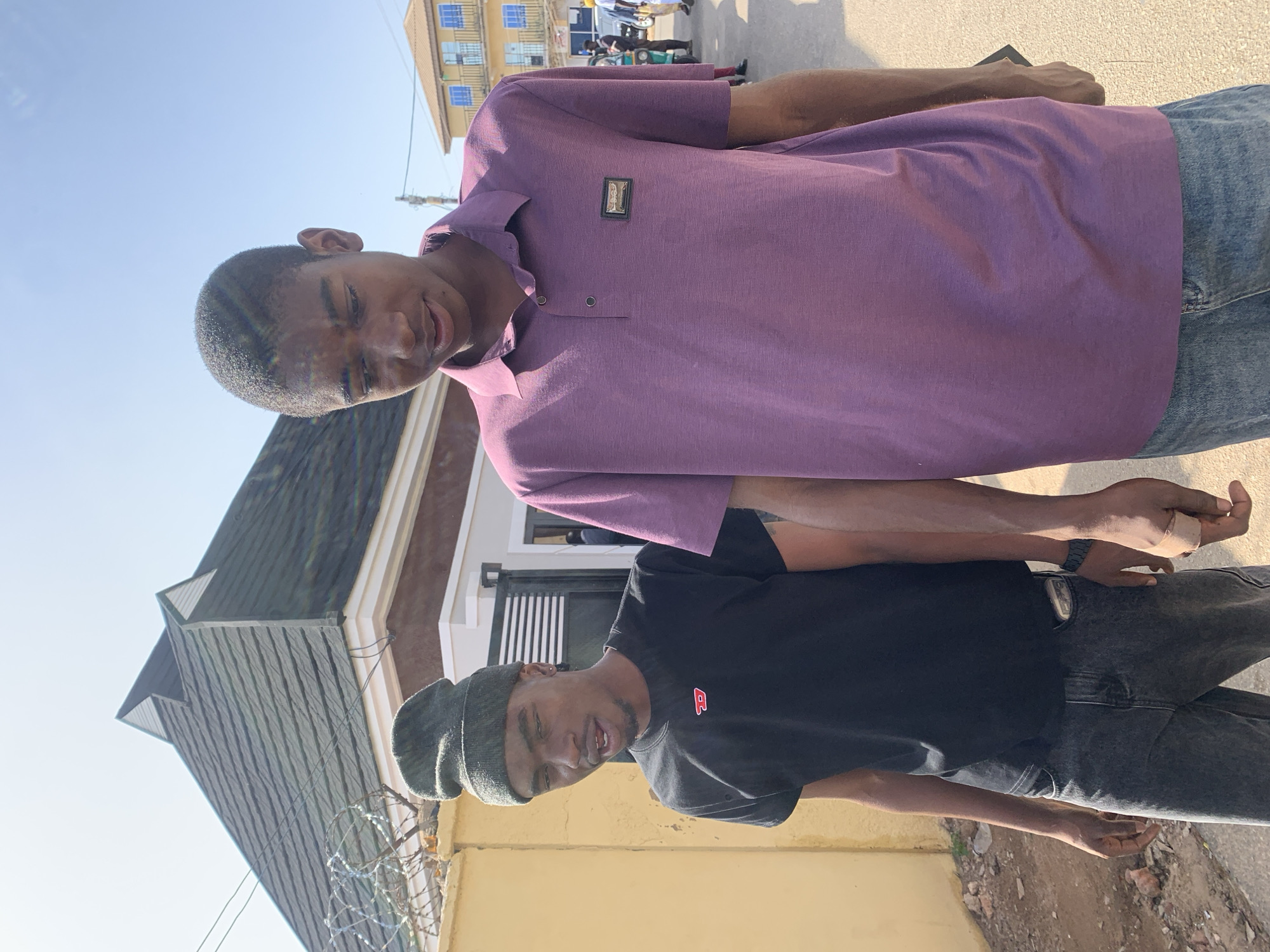 Two young African men are standing together. One is wearing a purple polo shirt and jeans, while the other is wearing a black t-shirt, grey jeans, and a dark beanie. They are positioned in front of a building with a dark grey roof. A yellow wall, topped with barbed wire, is visible in the foreground. The scene is located in Jos, Nigeria, evidenced by the architectural style of the buildings and the attire of the individuals. In the background, other people and vehicles can be observed.