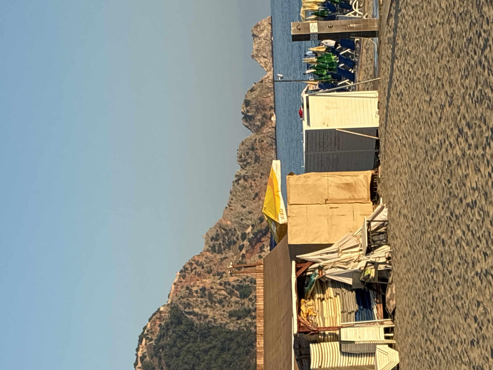 The image shows a beach scene in Alanya, Turkey. In the foreground, there is a sandy beach with several beach chairs and umbrellas set up. Behind the chairs, there are a few small structures that appear to be storage sheds or changing rooms. One shed is made of wood and covered in canvas, with stacks of yellow and white cushions inside. Another shed is made of light-colored wood. In the background, there are rocky mountains with vegetation and the blue sea with a few small islands in the distance. The sky is clear and blue. The sun appears to be setting or rising, casting long shadows across the sand.