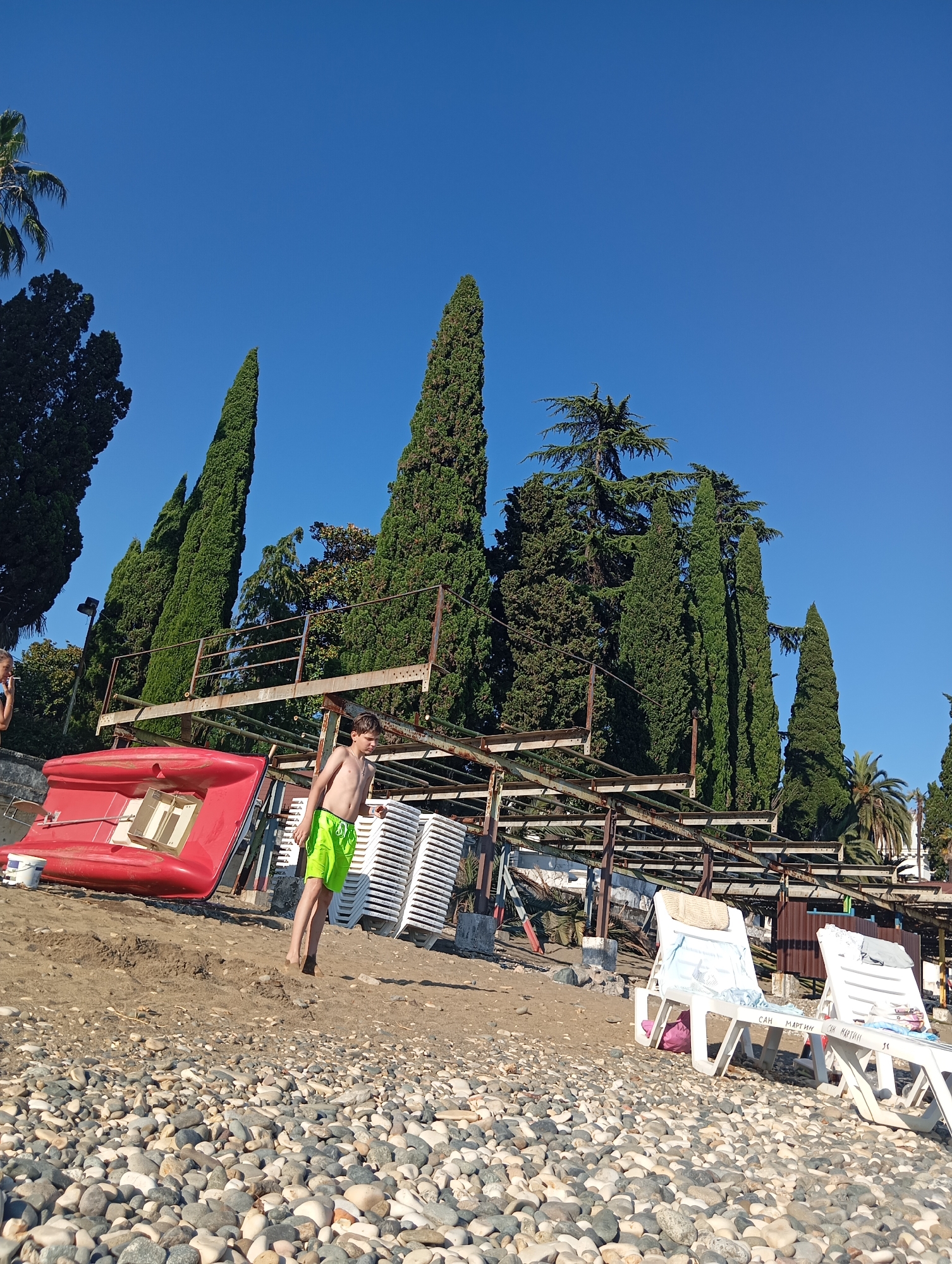 A young boy in green swim trunks stands on a pebble beach next to a red watercraft. Behind him are stacked white lounge chairs, a metal structure with rusted beams, and tall cypress trees. The sky is clear and blue. This scene appears to be at a beach resort in Sokhumi, Abkhazia, likely near a waterfront recreational area.
