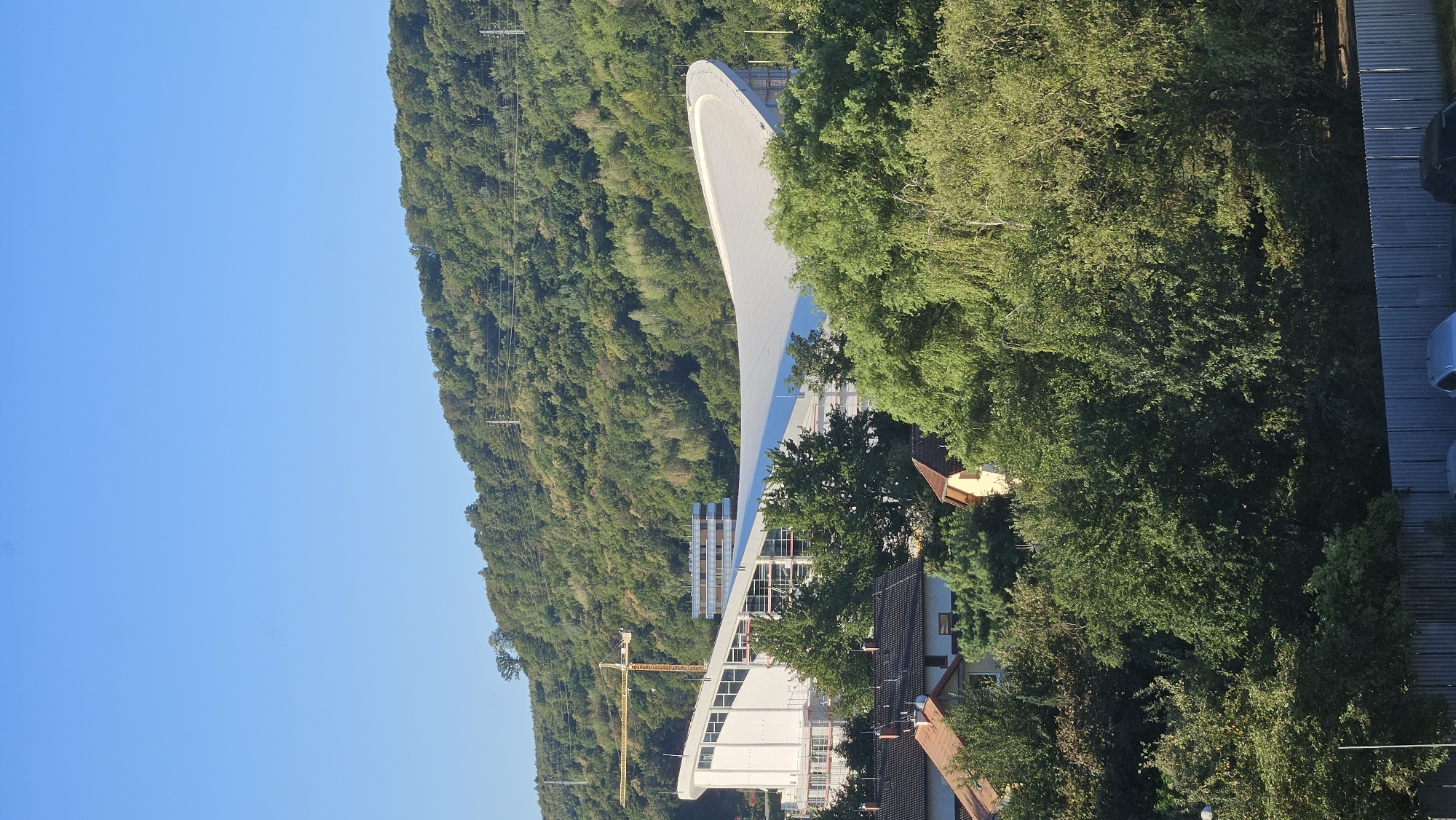The image shows a construction site with a large, modern building featuring a curved, white roof. A yellow crane is visible to the left of the building, indicating ongoing construction. Several smaller buildings with red-tiled roofs are in the foreground, suggesting a residential area. The background is dominated by a lush, green hillside covered in trees, under a clear blue sky. Electrical pylons are visible on the hillside. The scene appears to be a sunny day. The description provided in the prompt about the location being Prešov, Slovakia is not visually verifiable from the image alone.