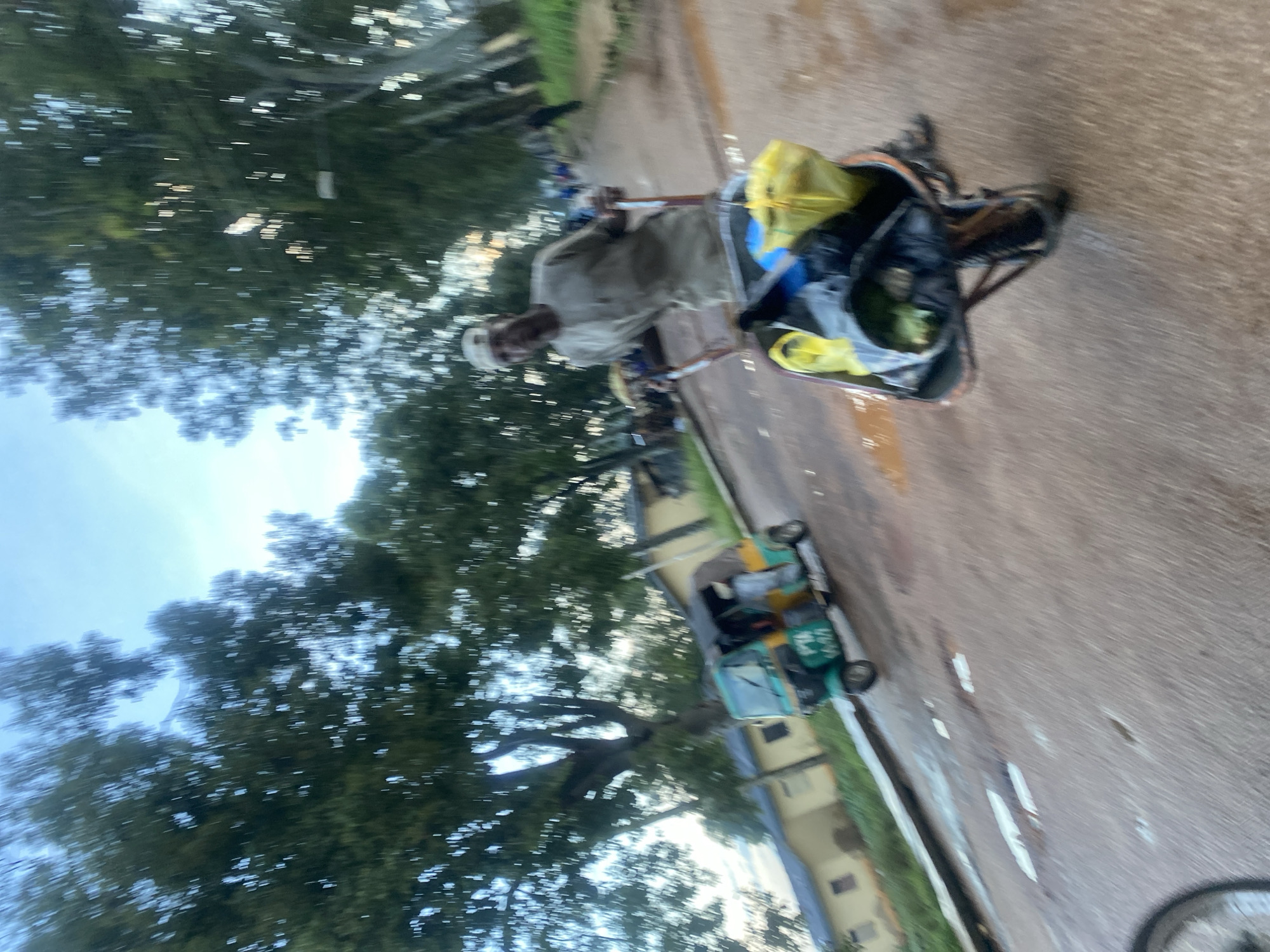 In Rafin Par, Nigeria, a man is pushing a wheelbarrow filled with bags down a wet, paved road. The wheelbarrow contains yellow, black, and gray bags. The man is wearing a light-colored traditional outfit and a white cap. Behind him, a teal and yellow three-wheeled vehicle, possibly a tuk-tuk, is driving on the road. The scene is bordered by trees on the left and buildings in the background. The sky is partly cloudy.
