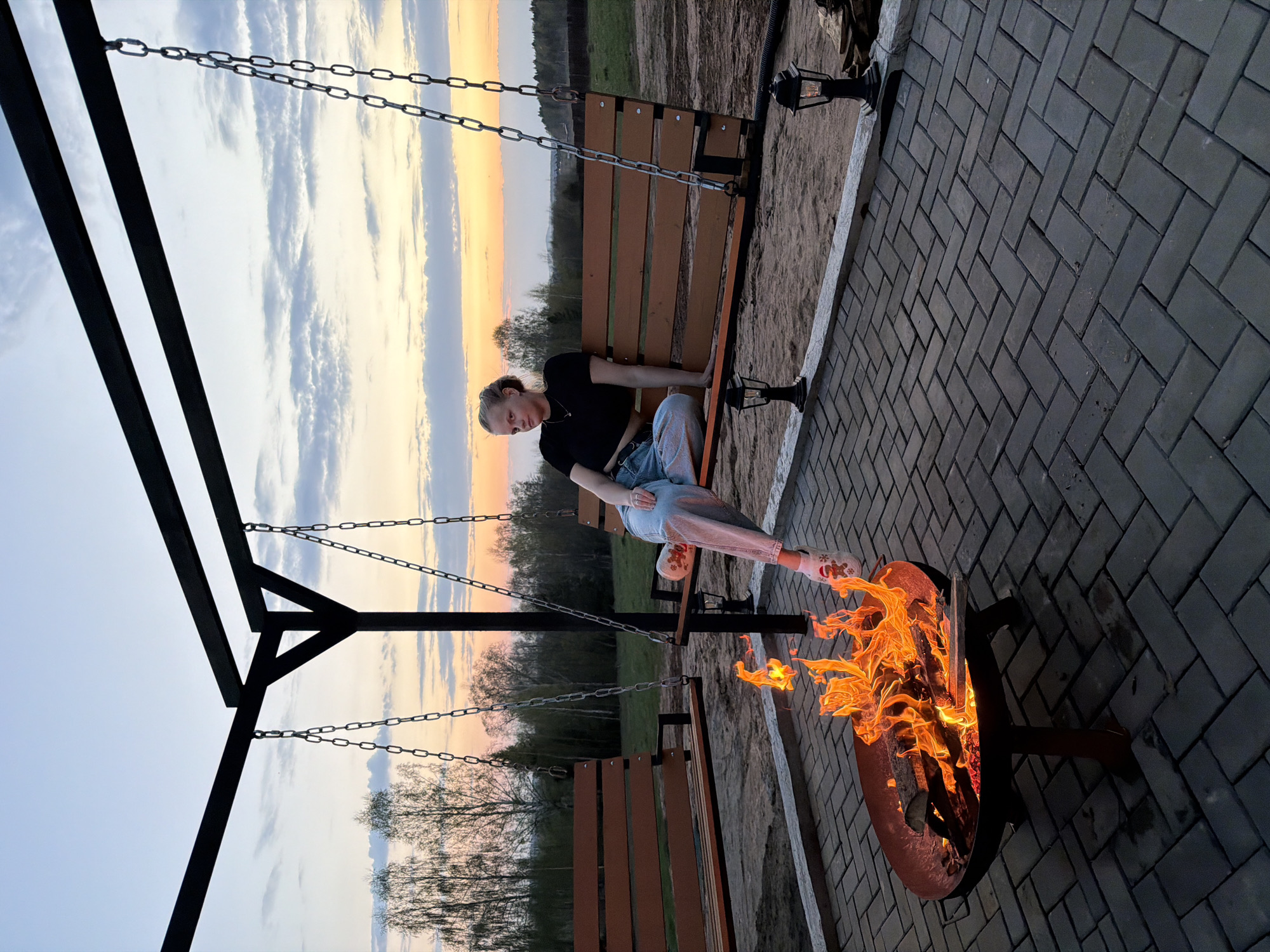 The scene shows a woman sitting on a wooden swing bench near a fire pit in an outdoor space. The paving is dark gray rectangular blocks. The sky transitions from light blue to orange and yellow at the horizon. A grassy field is visible in the background with trees. The woman wears a black shirt and light blue jeans with patterned white shoes and rests her feet near the fire pit. The location might be Kamennoye, Russia.