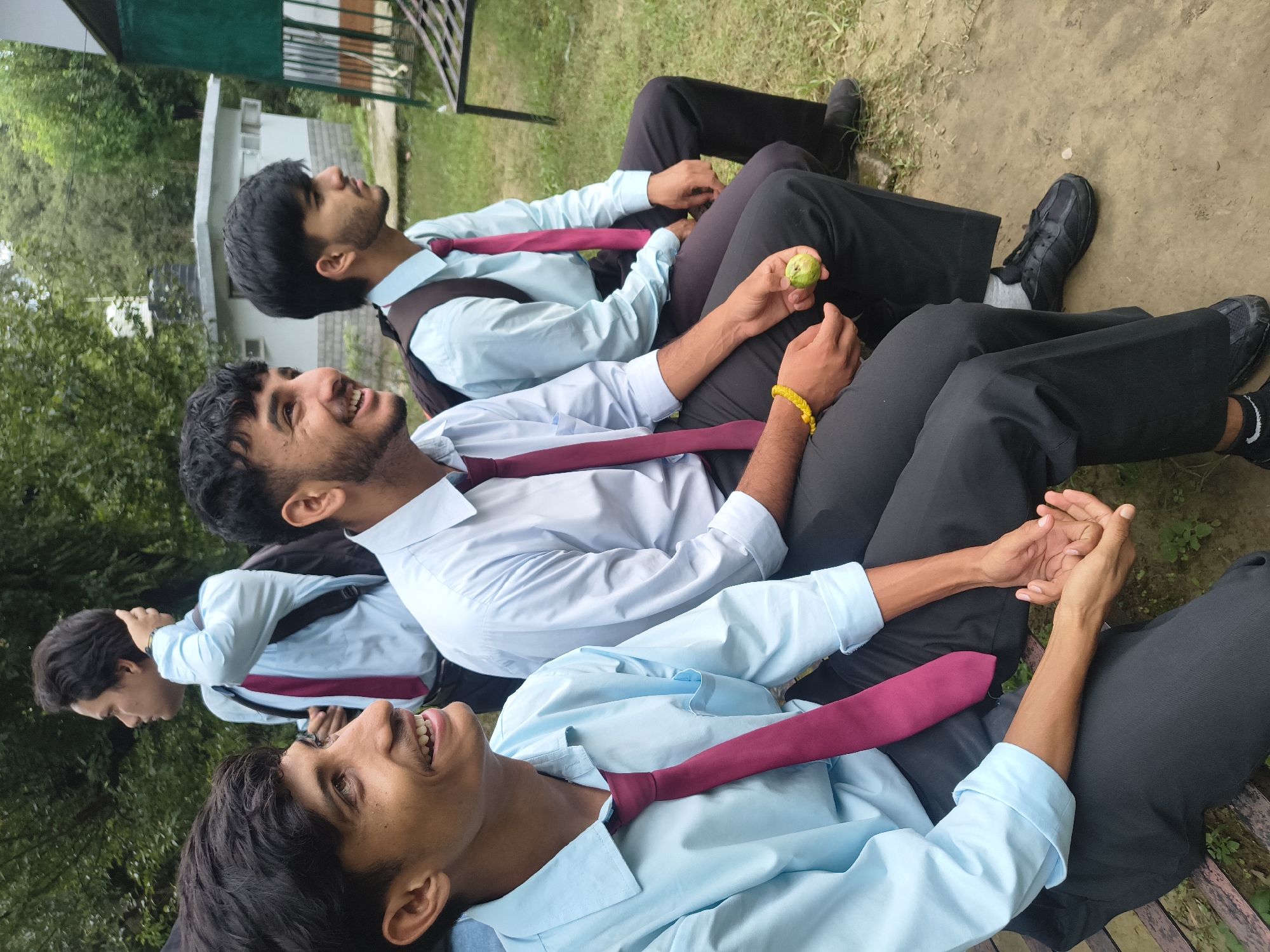 Four young men, presumably students, are seated on a bench outdoors. They are all wearing light blue collared shirts and dark ties. The man in the foreground on the left is smiling and looking to his left. He has dark hair and is wearing a light blue shirt and black trousers. To his right, another young man is also looking to his left with a smile. He has dark hair and a beard and is wearing a similar uniform. Further to the right, a third man, also with dark hair and a beard, is looking away from the camera. The fourth man is partially visible in the background, also wearing the same uniform. The men are seated on what appears to be a wooden bench. The environment is outdoors with trees and greenery visible in the background. There is a small white building with a darker roof behind the men and a dark green structure to the far right. The ground beneath them is a mixture of dirt and grass. The scene appears to be a casual gathering, possibly at a school or educational institution. This setting is consistent with the city of Dharamshala, India, known for its educational institutions and natural beauty.