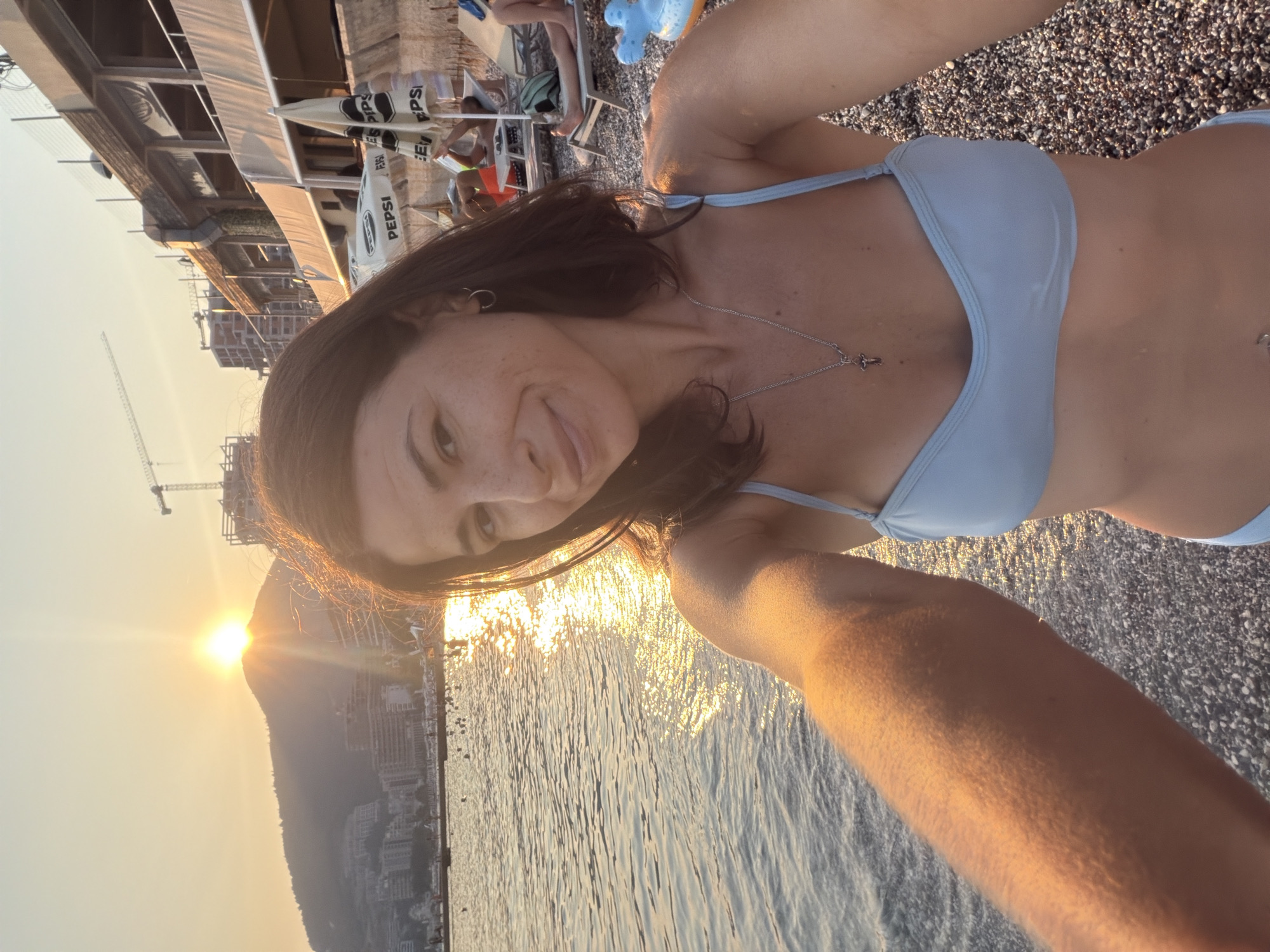A woman is taking a selfie on a pebble beach in Budva, Montenegro. The sun is setting over a mountain range in the background, casting a warm glow on the water and the scene. The woman is wearing a light blue bikini and a silver necklace with a cross pendant. She has a pierced belly button and small hoop earrings. In the background, there are several beach chairs, umbrellas, and people relaxing. A building under construction with cranes is visible on the hillside. The sea is calm with small waves.