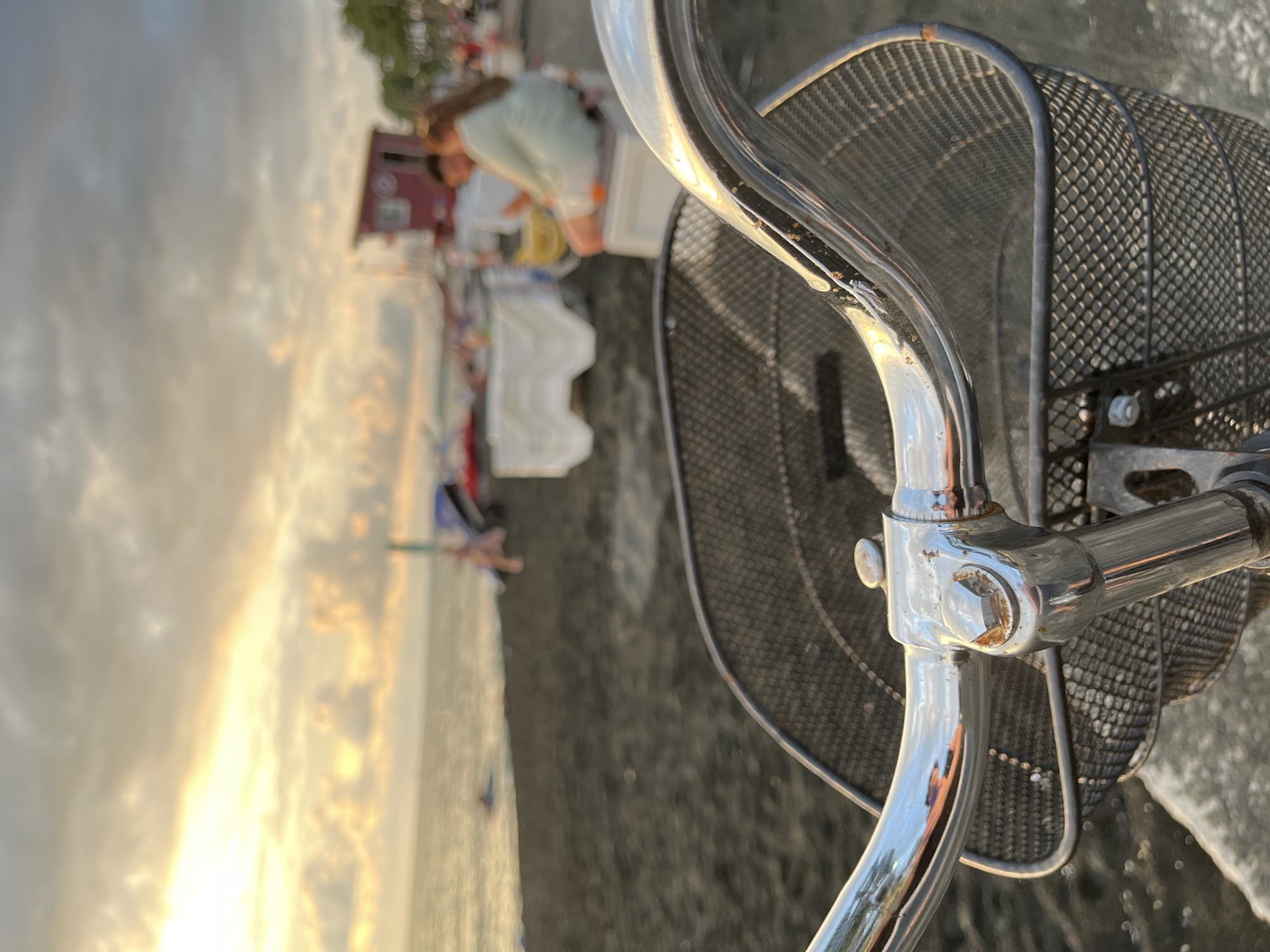 The image shows a close-up of a bicycle's handlebars and front basket, positioned on a black sand beach. In the background, the sea is visible, with a person swimming in the distance. Further inland, there are several people and beach amenities, including white chairs and tables, and a red lifeguard tower. The sky is cloudy with patches of golden sunlight, suggesting it is either sunrise or sunset. The overall scene depicts a relaxed atmosphere on the Ureki beach, known for its magnetic sand.