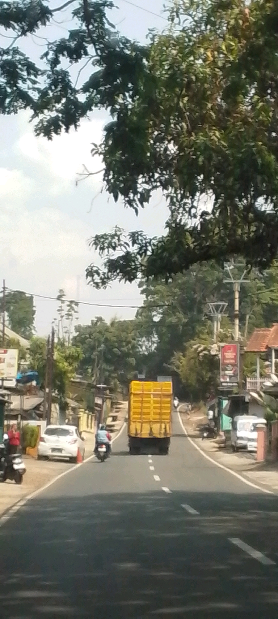 In Susukan, Indonesia, a narrow, asphalt road winds through a semi-urban environment with lush vegetation. The road is bordered by buildings and trees, suggesting a semi-rural or suburban setting. A bright yellow truck with a closed trailer is the dominant vehicle in the frame, traveling away from the viewer. On the left side of the road, a white car is parked near the curb, accompanied by a traffic cone. Further left, a motorcycle is parked. A person in a red shirt stands near the parked motorcycle, and another person is partially visible on a motorcycle in the distance. On the right side of the road, a white van is parked in front of a building with pink walls. The sky is overcast, with a mixture of clouds and sunlight breaking through. Overhead, large trees with dense foliage partially obscure the sky. Electrical poles and wires are visible along the roadside, indicating infrastructure. The overall scene depicts a typical day on a road in Susukan, with traffic and daily life unfolding.