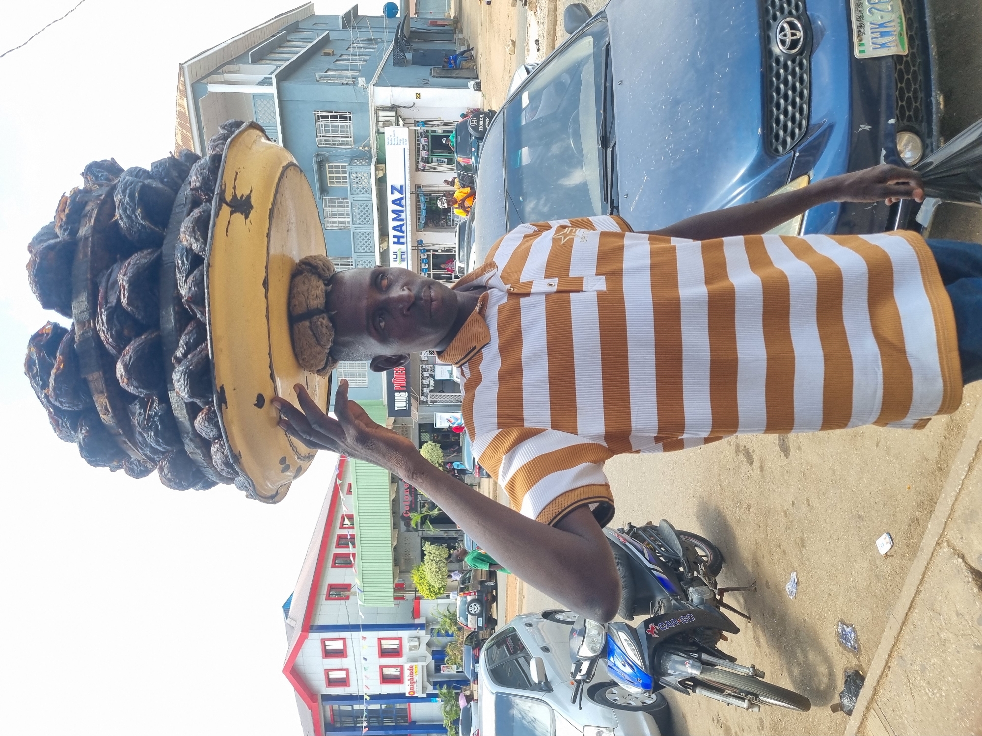 A man balances a yellow container piled high with kola nuts on his head on a street in Jos, Nigeria. He wears a striped white and brown polo shirt and looks directly at the camera. Cars and buildings line the street in the background. A blue motorcycle is parked near him. License plates are visible on the cars in the background.