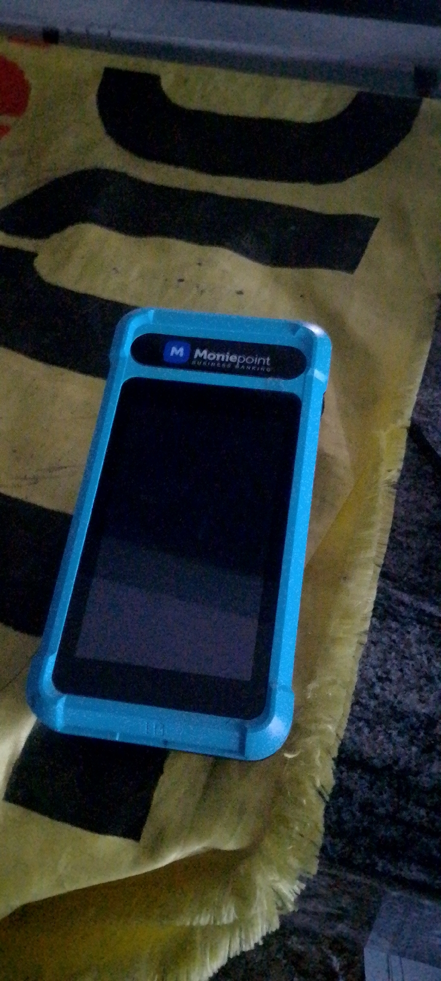A bright blue, rugged case is fitted around a black rectangle, likely a point-of-sale device or tablet. The case has a cutout at the top which reveals a dark blue rectangle with the "M Moniepoint BUSINESS BANKING" logo in white text. The device is lying on a fuzzy, yellow and black striped surface. The background is dark and out of focus.