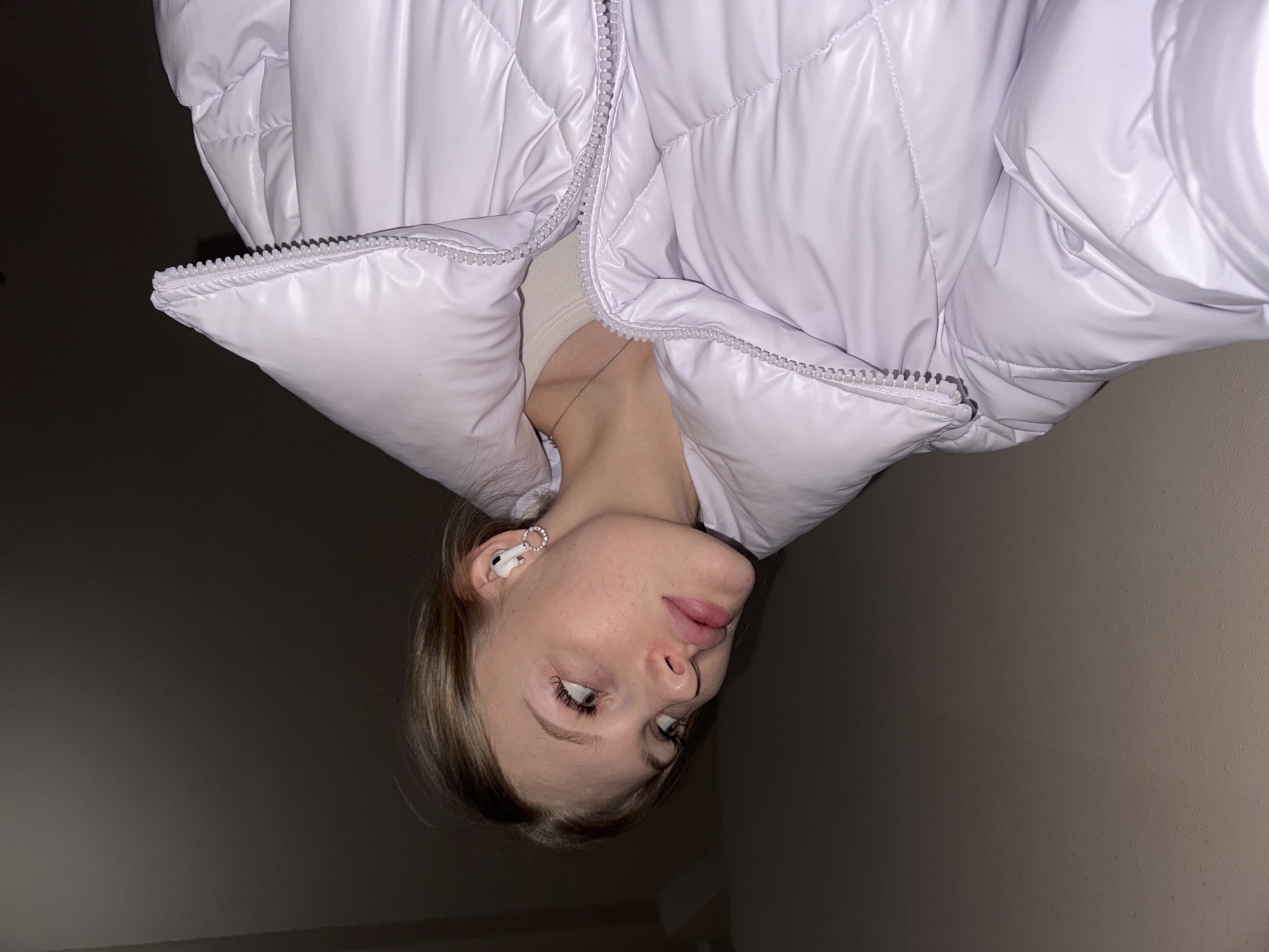 The image, originally rotated 180 degrees, features a young woman in an indoor setting, identified as Perm, Russia. She is shown from the chest up, wearing a glossy, light lavender (or pale purple) puffer jacket with a quilted pattern and a visible zipper. A light-colored undershirt, possibly cream or white, and a thin silver-toned necklace are visible beneath the unzipped jacket.
Her face is in focus, displaying a neutral to slightly contemplative expression with her lips subtly parted. Her light-colored hair, appearing blonde or light brown, is styled casually around her face. She has AirPods in her left ear, which are embellished with a circular earring featuring small, clear decorative stones. Her eyes are light-colored and gaze slightly upwards and to the side (when the image is viewed upright).
The background consists of a plain, dark wall, likely dark grey or brown, which contrasts with the light color of her jacket. The lighting appears to be artificial, possibly from an overhead source, casting some subtle shadows and highlighting the shiny texture of the jacket. The image captures a casual moment or a self-portrait. There is no visible text.