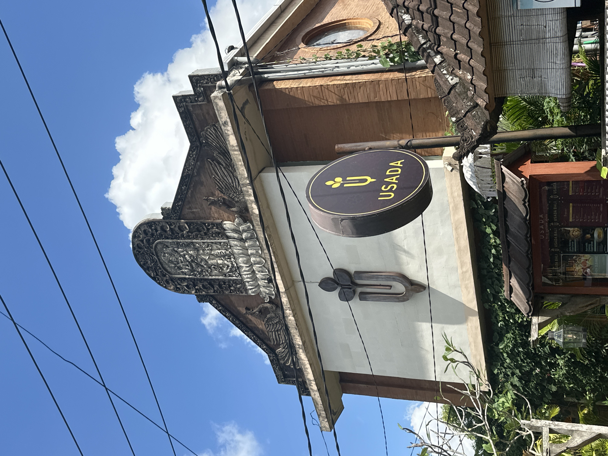 A low-angle outdoor medium shot captures the facade of the "USADA" building, a health and wellness center, in Ubud, Indonesia. The building features Balinese architectural elements, including intricately carved stonework on the upper part of the facade, depicting figures and motifs. A large circular sign with the text "USADA" and a stylized plant logo hangs from a pole in front of the building. The building is constructed with a combination of white plaster and reddish-brown brick. Lush green foliage covers the lower portion of the building, and electrical wires are visible crisscrossing the bright blue sky, which is dotted with white clouds. A tiled roof structure with decorative elements is visible in the foreground to the right, partially obscuring a window on the building. A small wooden structure with a menu board is present to the left of the tiled roof. The scene is bathed in natural daylight, suggesting it was taken during the daytime.