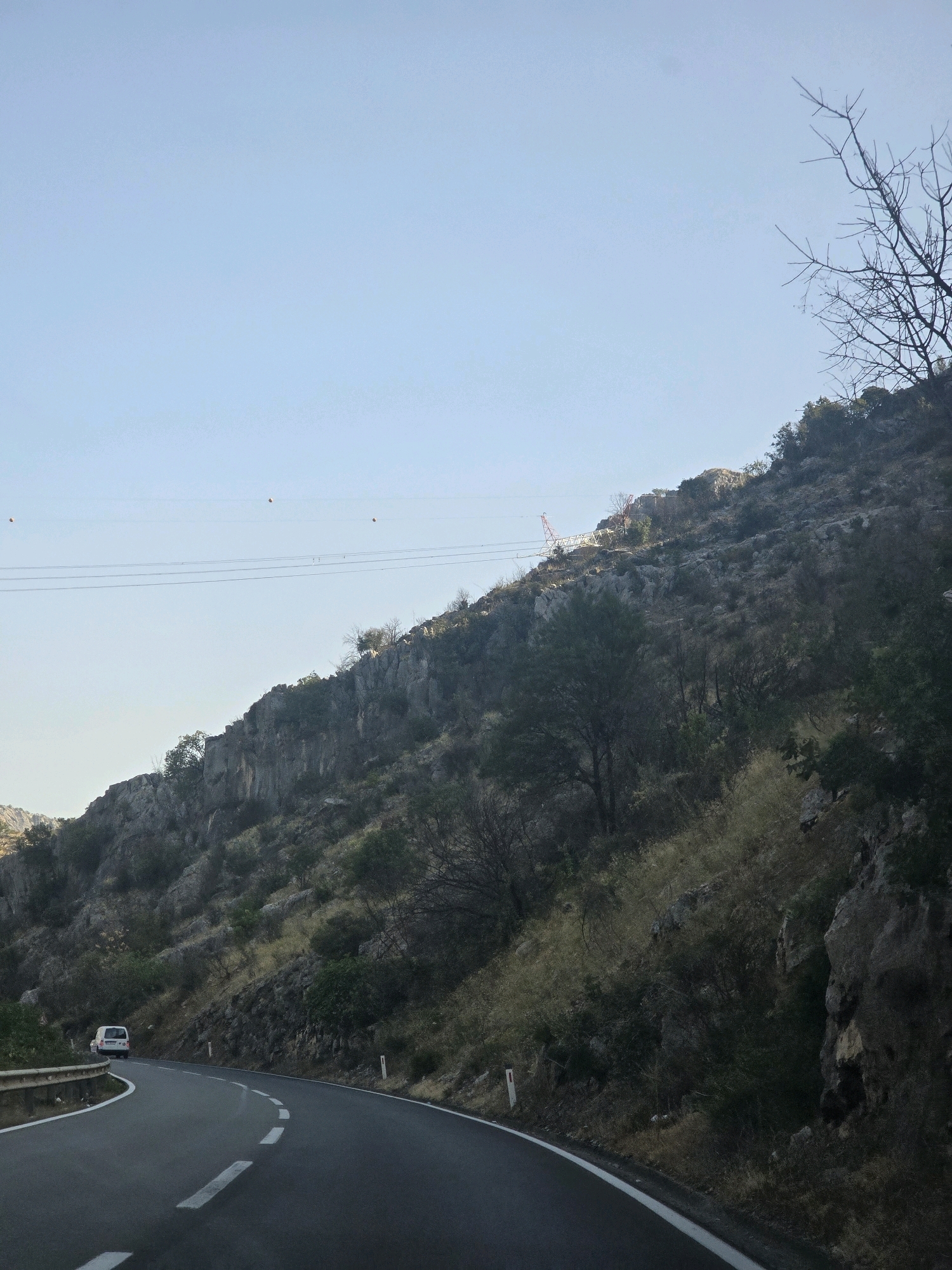 The image depicts a winding road in a mountainous, semi-arid region, likely in the vicinity of Podgorica, Montenegro. The road curves to the left, with a white guardrail on its left side. A white van is visible driving on the road. The terrain flanking the road is characterized by rocky slopes covered with sparse vegetation, including dry grasses and small trees. In the background, power lines stretch across the sky, supported by poles, and a large red and white transmission tower is visible on the top of a hill. The sky is clear and pale blue, suggesting a sunny day. The overall impression is one of a rural or semi-rural mountain landscape.