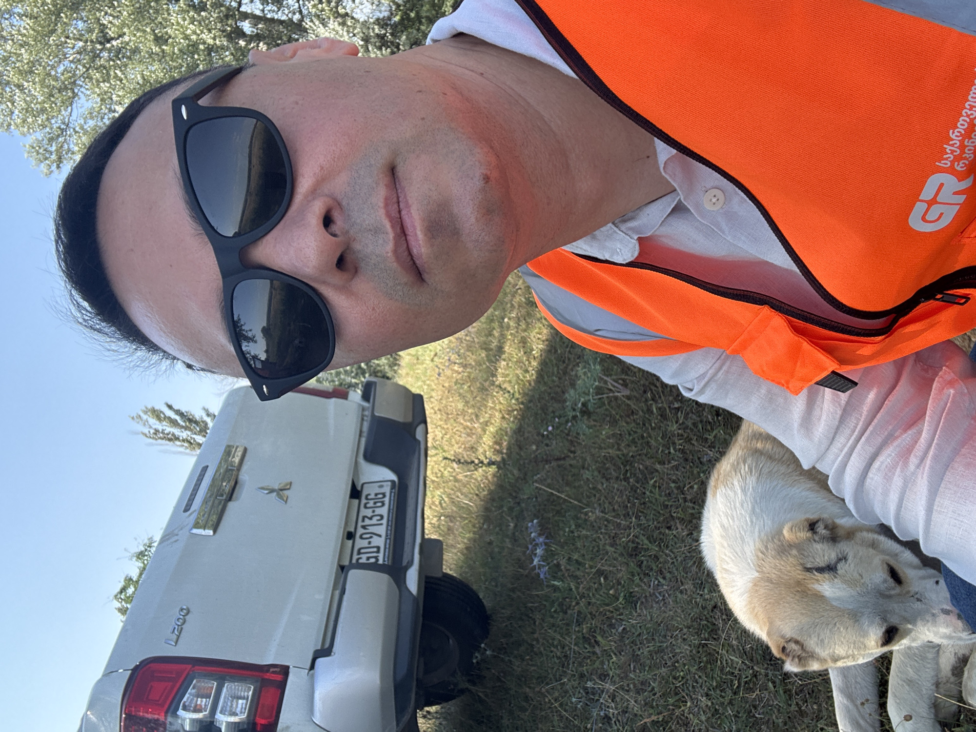 A man wearing sunglasses and an orange safety vest is taking a selfie outdoors. To his left, a white Mitsubishi L200 pickup truck is parked. The license plate is visible and reads "GD-913-GG". A large dog, light brown and white, is lying on the grass next to the man. The background shows dry grass, some trees, and a clear blue sky, suggesting a rural or semi-rural setting in Kvemo Khvedureti, Georgia. The overall scene appears to be a casual moment outdoors, possibly during a work-related activity given the safety vest.