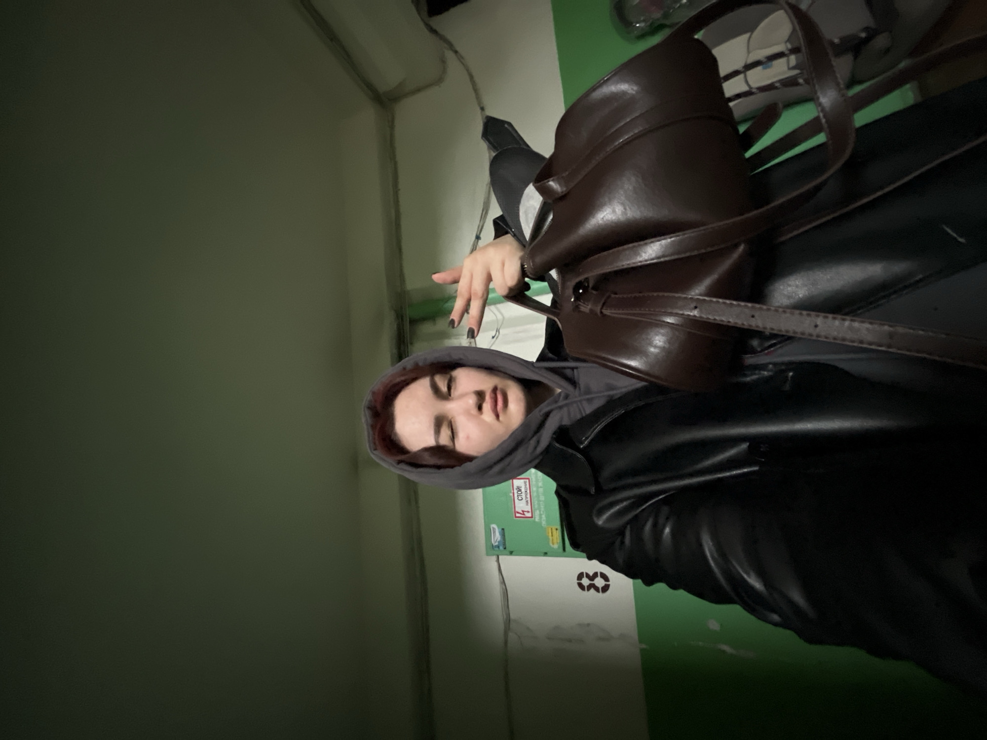 A person wearing a gray hoodie and a black leather jacket is standing in a hallway with green and white walls. The person has dark hair and is holding a brown leather bag. The location may be in the city of Neftekamsk, Russia.