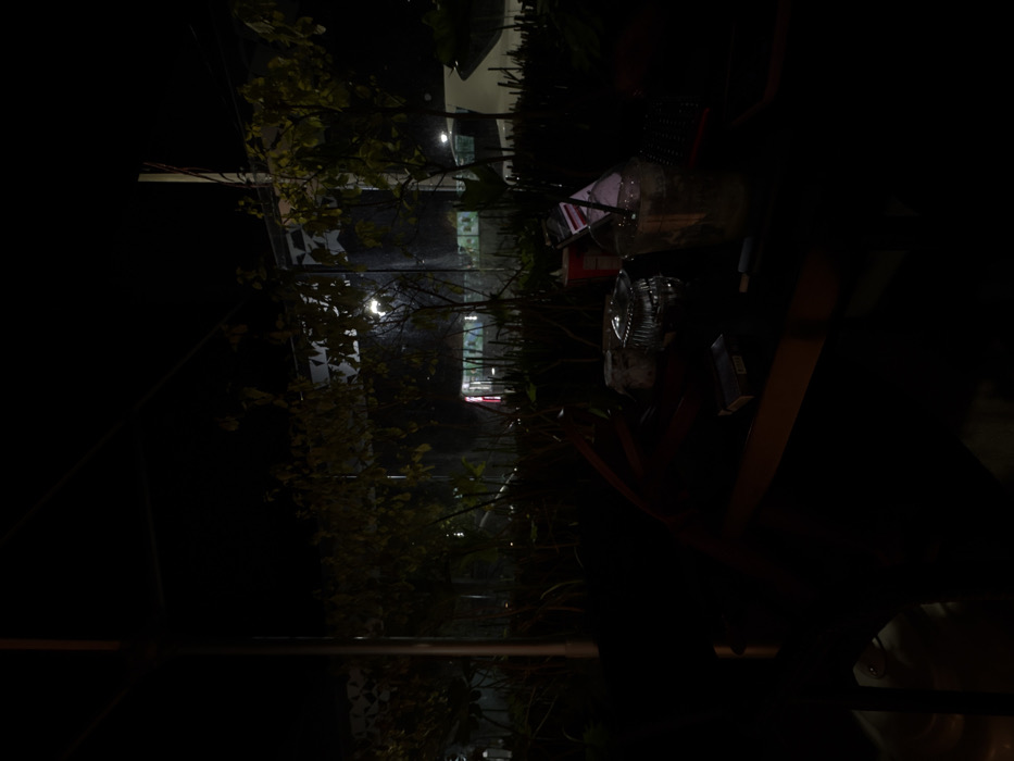 A nighttime shot reveals a blurred table with various items, including a clear plastic bottle, a can, and a pack of cigarettes. Outdoor vegetation frames the scene with glimpses of distant buildings and streetlights, creating a depth perspective. The overall ambiance is dark, with the objects on the table being partially illuminated and blurry. The image has been rotated and needs to be viewed sideways.