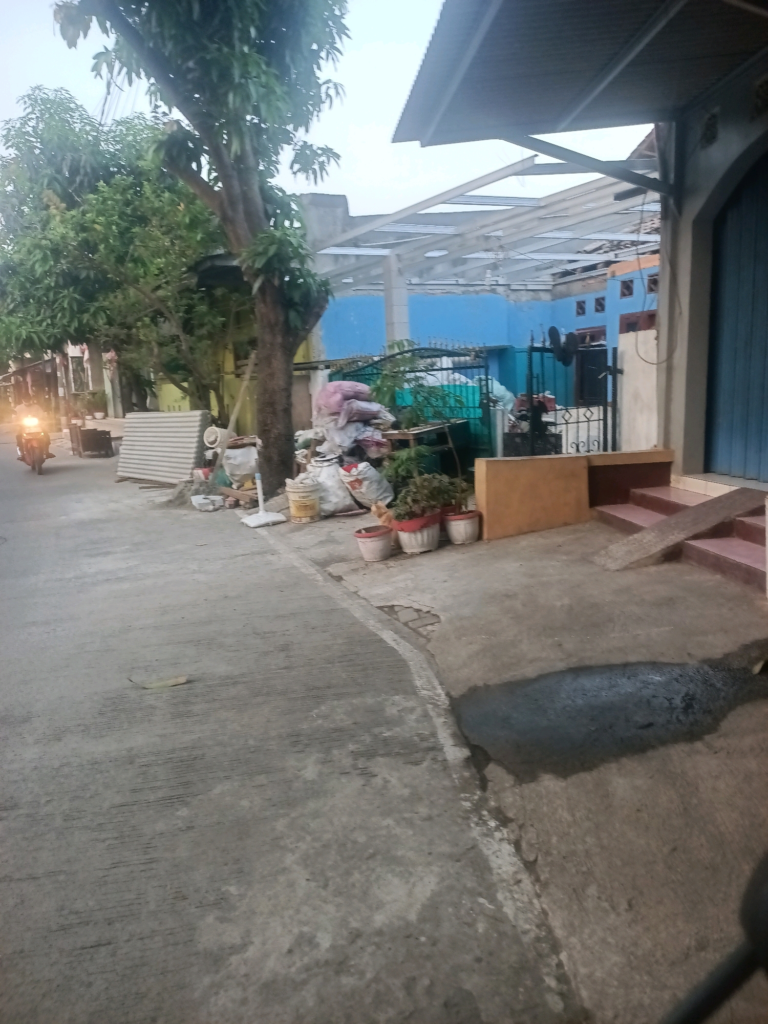 The image depicts a street scene in Telagaasih, Indonesia, with a focus on a roadside area cluttered with various objects. A motorcycle with its headlight illuminated is visible on the left side of the image, moving along a paved road. To the right of the road, there is a significant amount of debris and household items piled up, including large plastic bags filled with unknown contents, a yellow bucket, several potted plants, and what appears to be corrugated metal sheeting stacked vertically. Further to the right, a building with a blue facade and a metal roof structure is partially visible. There is a set of stairs leading up to the building, with a wooden ramp placed on one of the steps. The ground surface is concrete, with some patches of dark, wet-looking material. Vegetation, including trees and bushes, frames the scene on the left and in the background. The overall impression is one of an outdoor, possibly residential or commercial area, with a noticeable accumulation of discarded items.