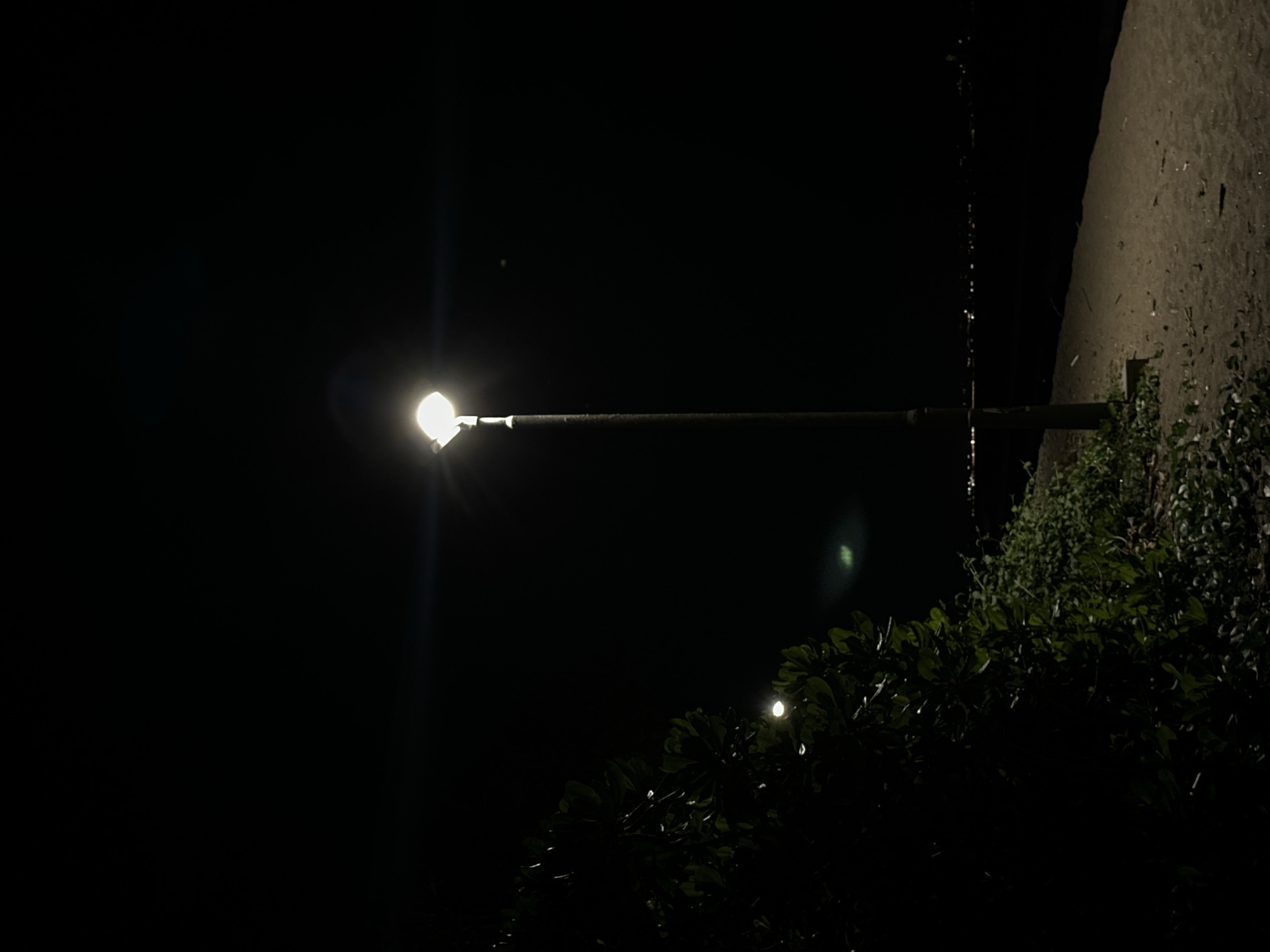 A street lamp illuminates a section of a wall and some bushes in North Kuta, Indonesia. The image is taken at night, with the majority of the frame obscured by darkness. The illuminated wall is textured, possibly concrete. Below the wall, bushes are visible, partially illuminated by the street lamp and another light source behind the bushes, on the lower left side of the image. The street lamp extends from the wall.