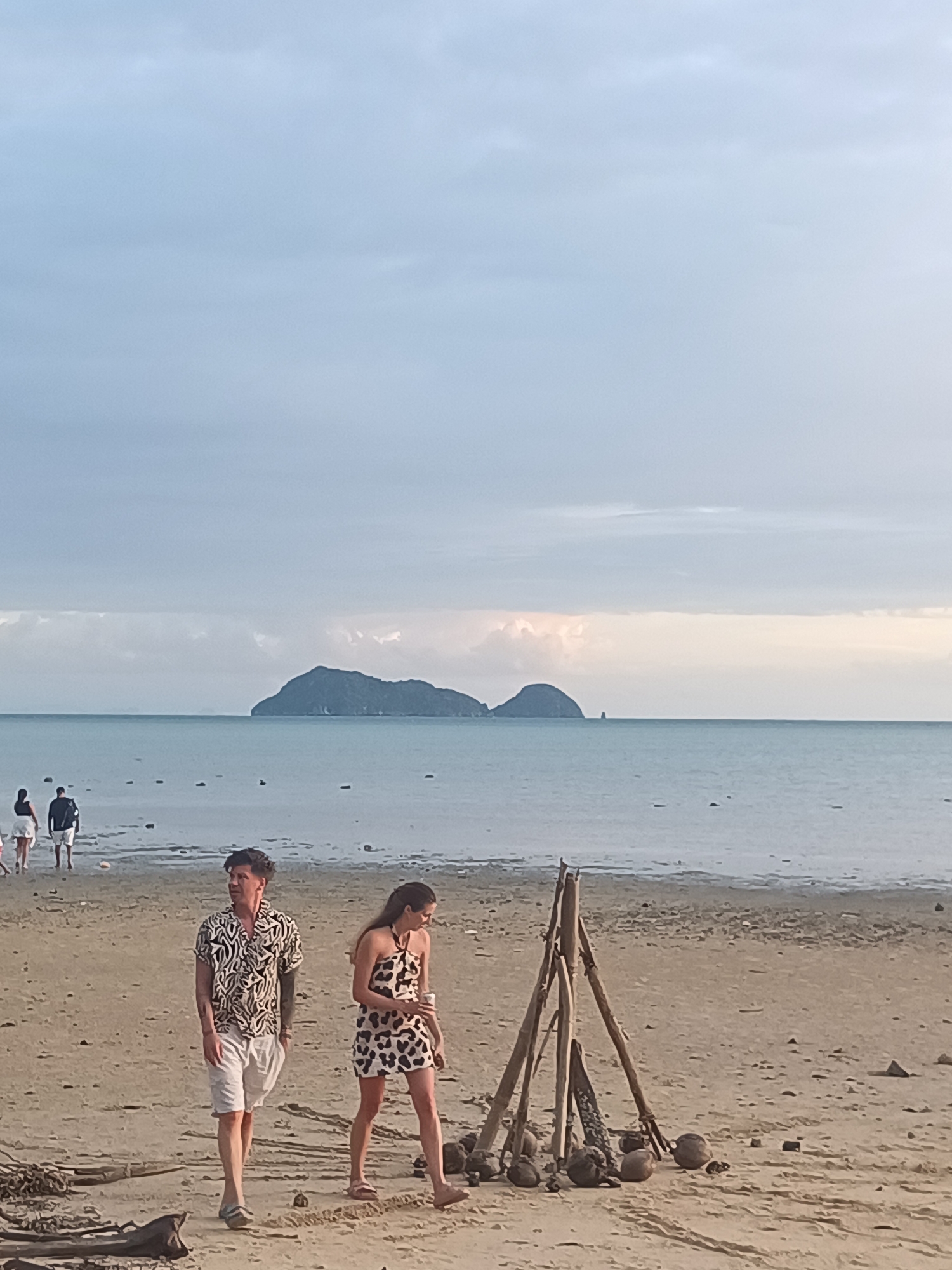 The image depicts a beach scene with a man and a woman walking on the sand in the foreground. The man is wearing a short-sleeved patterned shirt and white shorts, while the woman is wearing a patterned dress. Behind them, in the middle ground, several other people are walking near the water's edge. In the background, a body of water, likely the sea, is visible with islands in the distance under a cloudy sky. There is a makeshift structure of sticks and coconuts on the beach in the foreground. The overall environment is a beach in ตำบลเกาะพะงัน, Thailand, suggesting a tropical coastal setting.