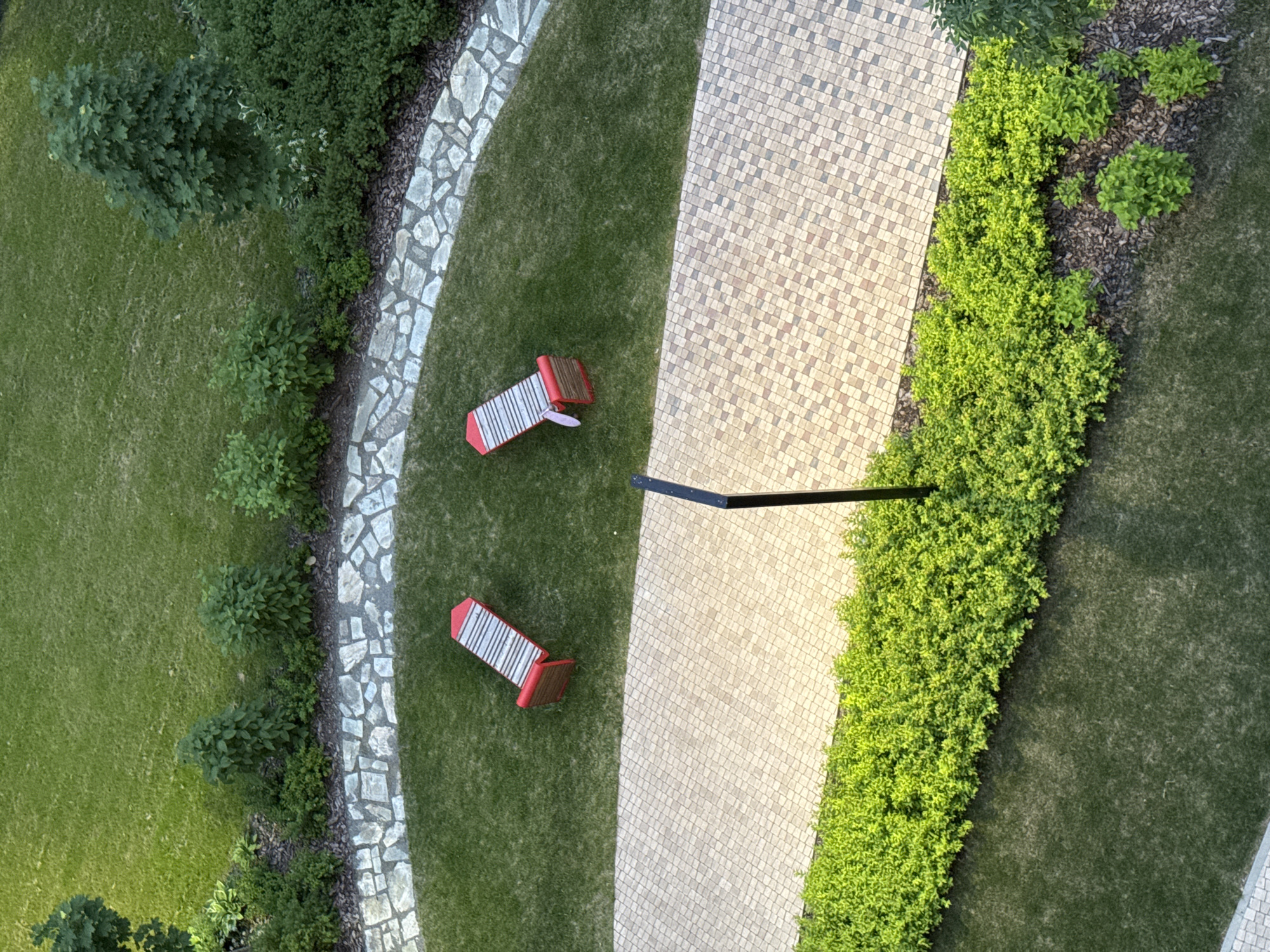 The image shows an aerial view of a park in Izhevsk, Russia. In the center of the frame are two red and white striped benches situated on a patch of green grass. The grass is bordered by a curved pathway made of light-colored flagstones. Beyond the flagstone path, to the right, is a paved area with a lamp post. A dense hedge of bright green foliage runs along the bottom of the paved area. Further to the left, the flagstone path curves around another grassy area with more vegetation. The overall scene depicts a well-maintained outdoor public space.