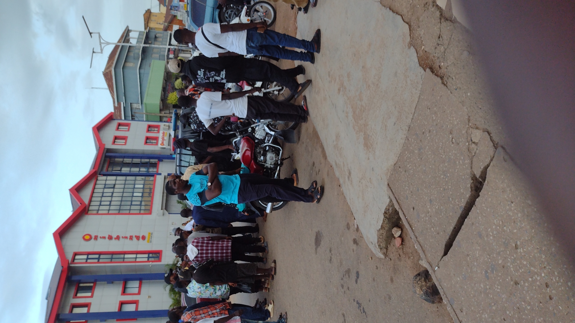 A group of men are gathered on a paved street in front of a modern, two-story building with a red roof and blue accents. The building has "Onighinde" signage visible. Several motorcycles are parked near the building, with one red motorcycle positioned prominently in the foreground. The men are casually dressed, some are standing and talking, while others appear to be observing. The sky is overcast, suggesting a cloudy day. The scene is likely located in Jos, Nigeria, given the context provided by the user.