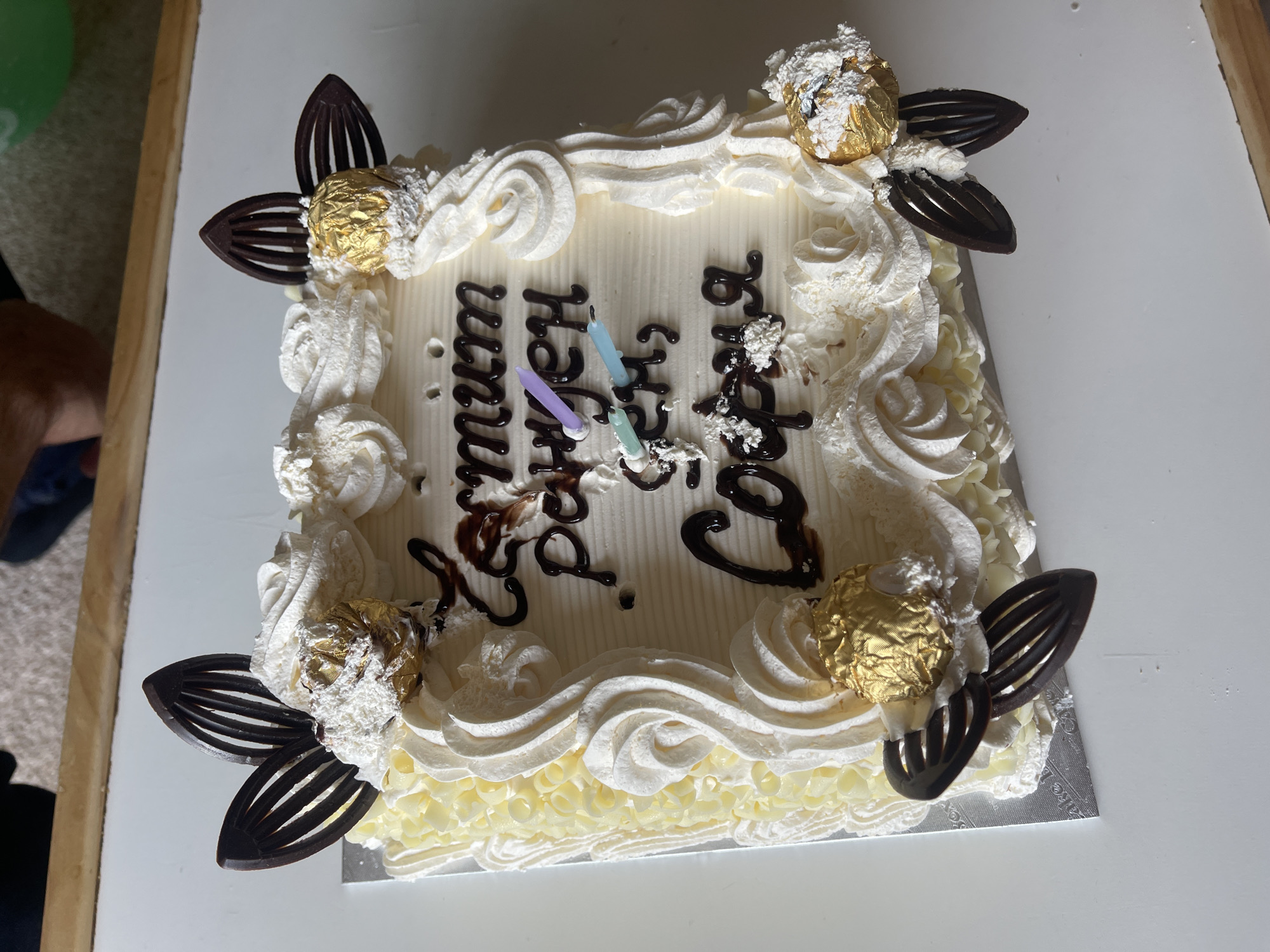 A square birthday cake, likely in a domestic setting. The cake has white frosting with decorative swirls along the top edges and a layer of smaller decorative elements along the bottom edge. Four Ferrero Rocher chocolates, each surrounded by a small nest of white frosting and four dark chocolate leaf decorations, adorn the corners of the cake. "Happy Birthday" is written in dark chocolate on the top of the cake, oriented vertically. Three birthday candles are placed in the center of the written message. Parts of a person and furniture are visible in the background. No specific location can be identified to be Maidstone.