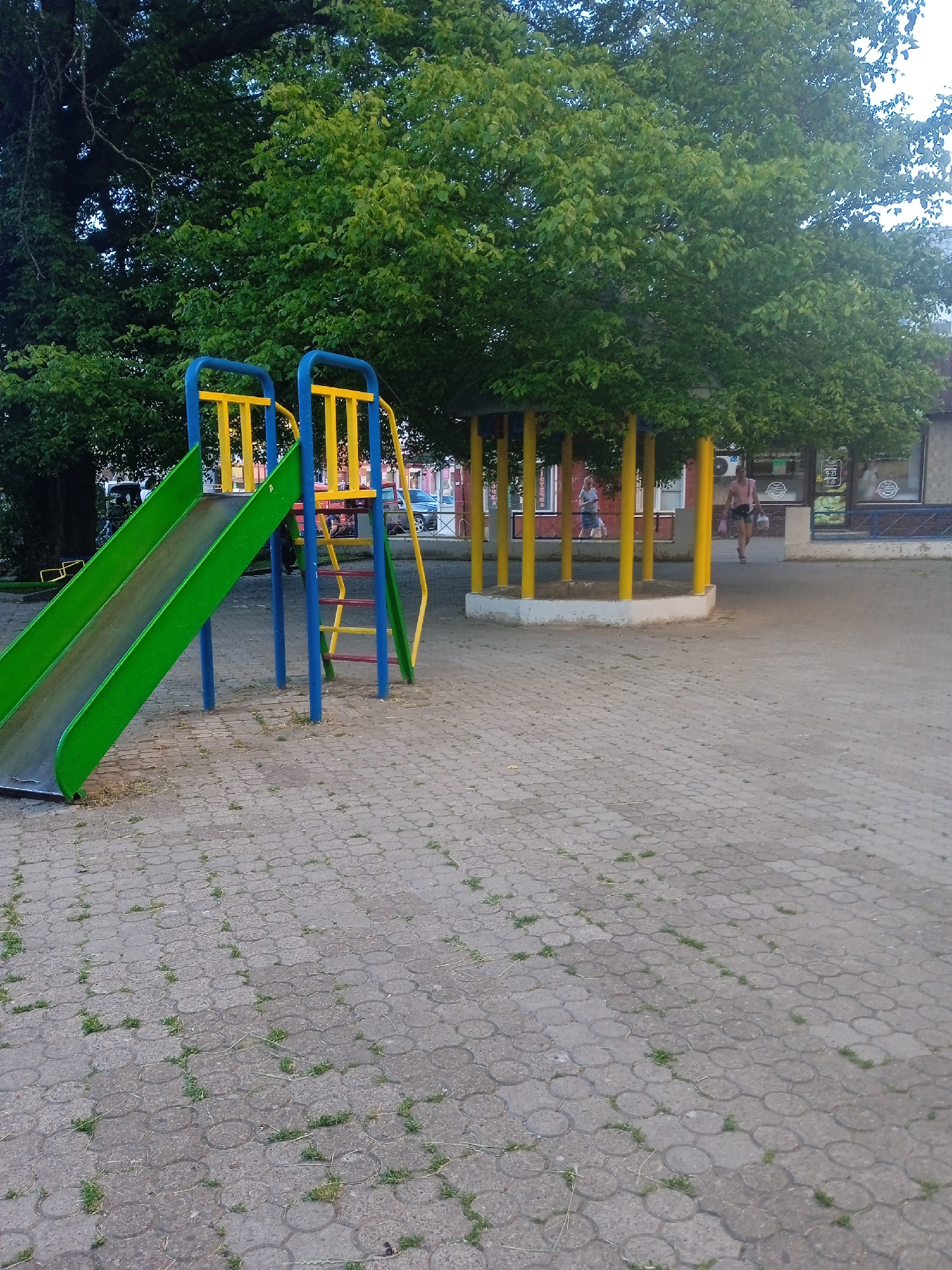 The image depicts an outdoor public space in Chernivtsi, Ukraine. In the foreground, a green and yellow playground slide with blue support poles is visible, set on a gray paver stone surface. Behind the slide, a large tree with dense green foliage dominates the background, casting shadows. To the right of the slide, a circular structure with yellow support columns and a white base is present, appearing to be part of an open-air shelter. In the middle ground, two individuals are walking. One, a person in a light pink top and dark shorts, is walking away from the camera, carrying a pink bag. Another person, wearing a light blue top and dark shorts, is walking towards the camera in the background. Buildings with storefronts are visible behind the sheltered area. The overall scene suggests a park or communal area in a city.