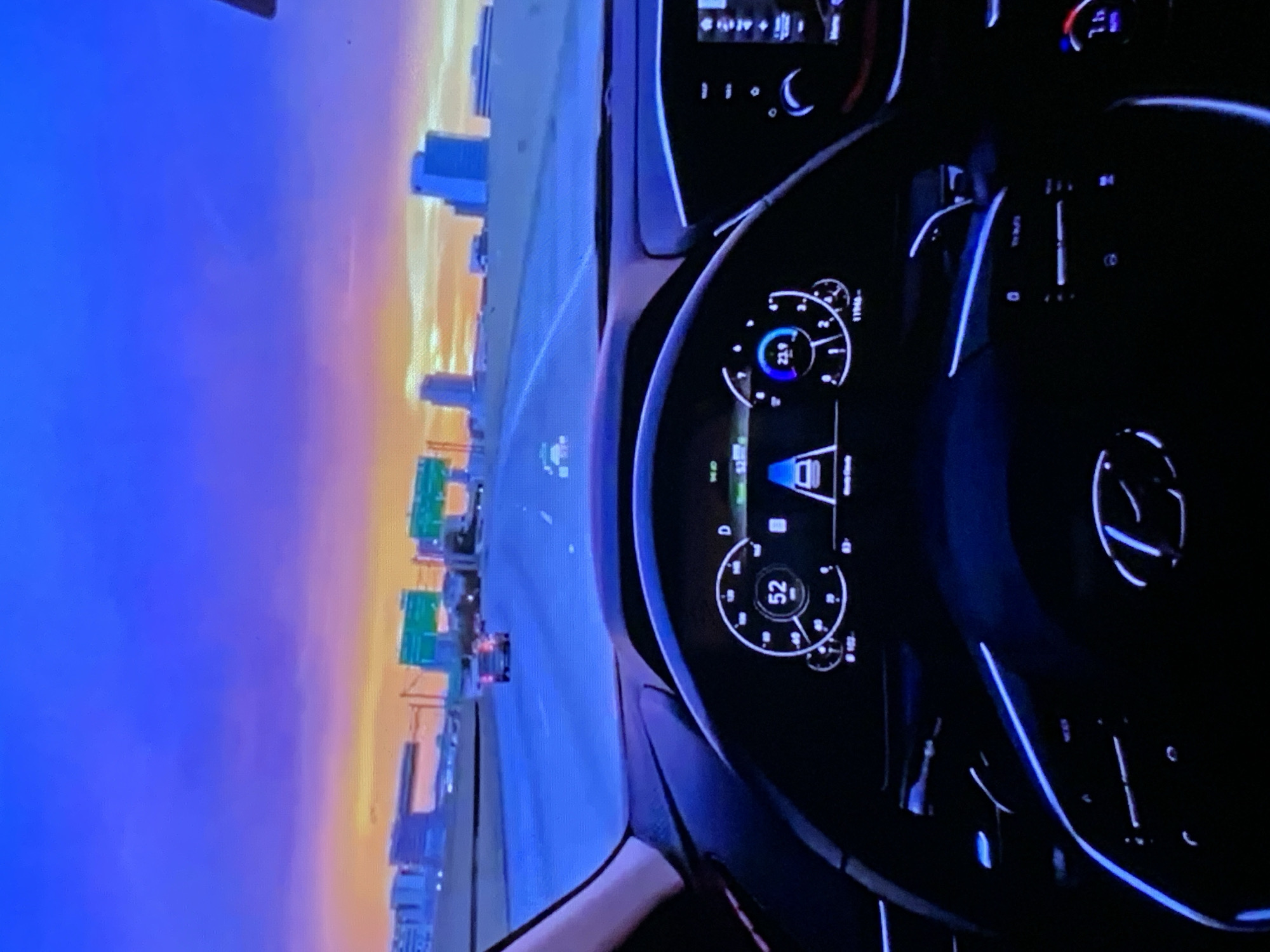 The image shows a view from the driver's seat of a car on a highway at dusk. The sky is a vibrant purple and orange, suggesting a sunset or sunrise. In the distance, a city skyline is visible with several tall buildings. The dashboard of the car is illuminated, showing a digital speedometer displaying 52 mph, and other indicators. The steering wheel, with the car's emblem, is also visible. The highway is multi-laned and there are other cars on the road. The overall scene depicts a drive through an urban area during twilight.